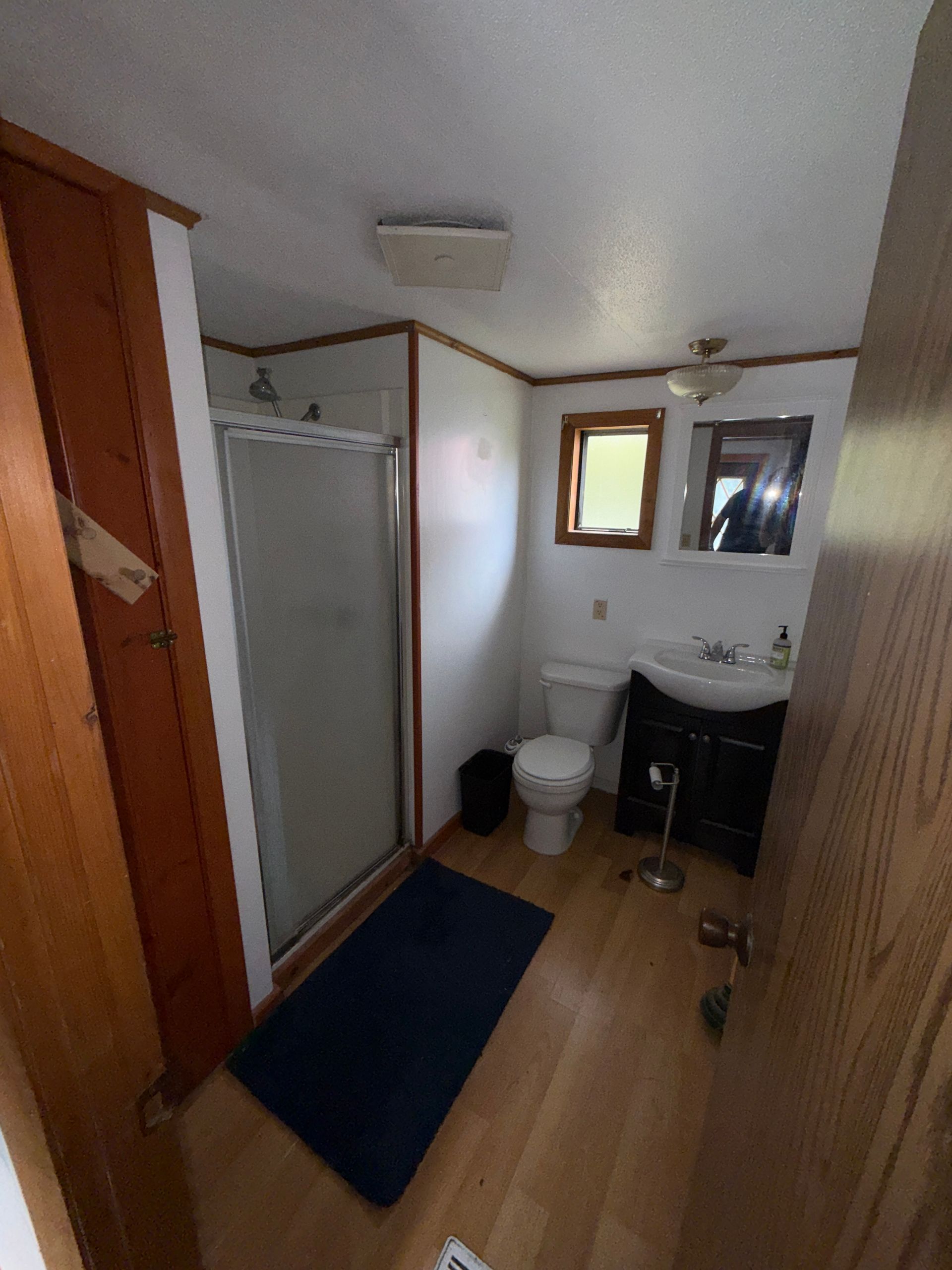 Small bathroom with shower, toilet, sink, dark rug, and mirror near a wood floor.