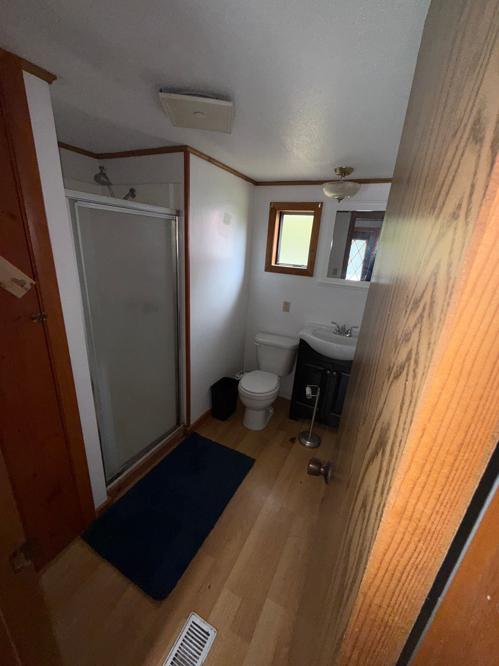 Small bathroom with shower, toilet, sink, window, and blue rug on wood floor.