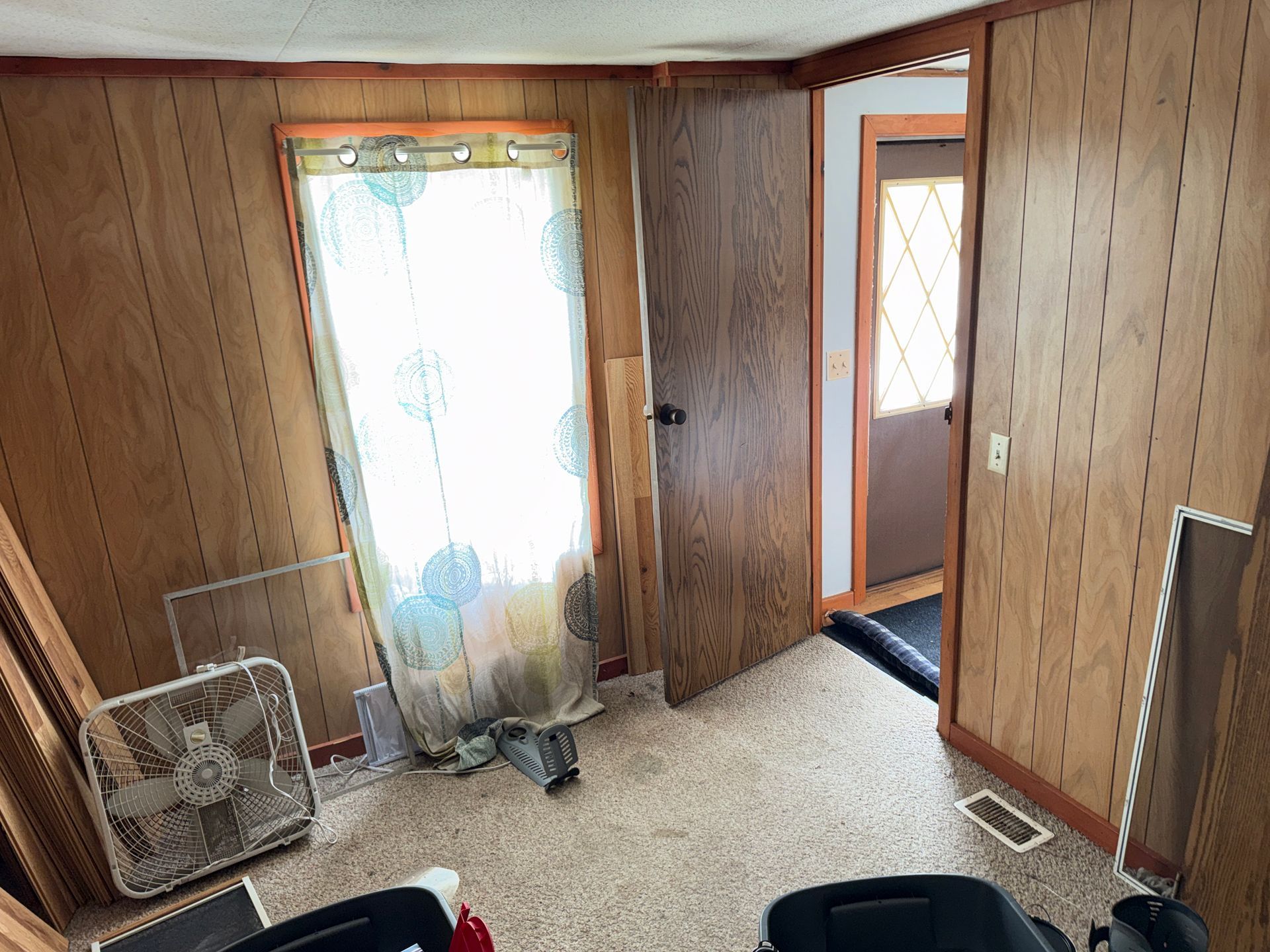 Wood-paneled room with carpet, a bright curtained window, open door, and a box fan on the floor.