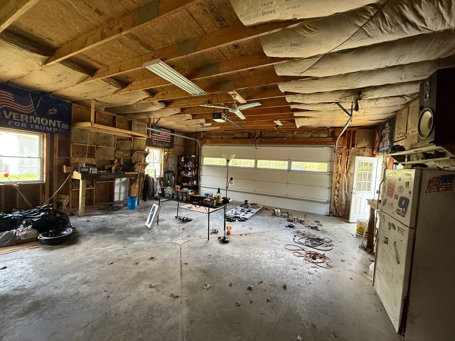 Garage with exposed insulation, tools, and a white refrigerator near the door