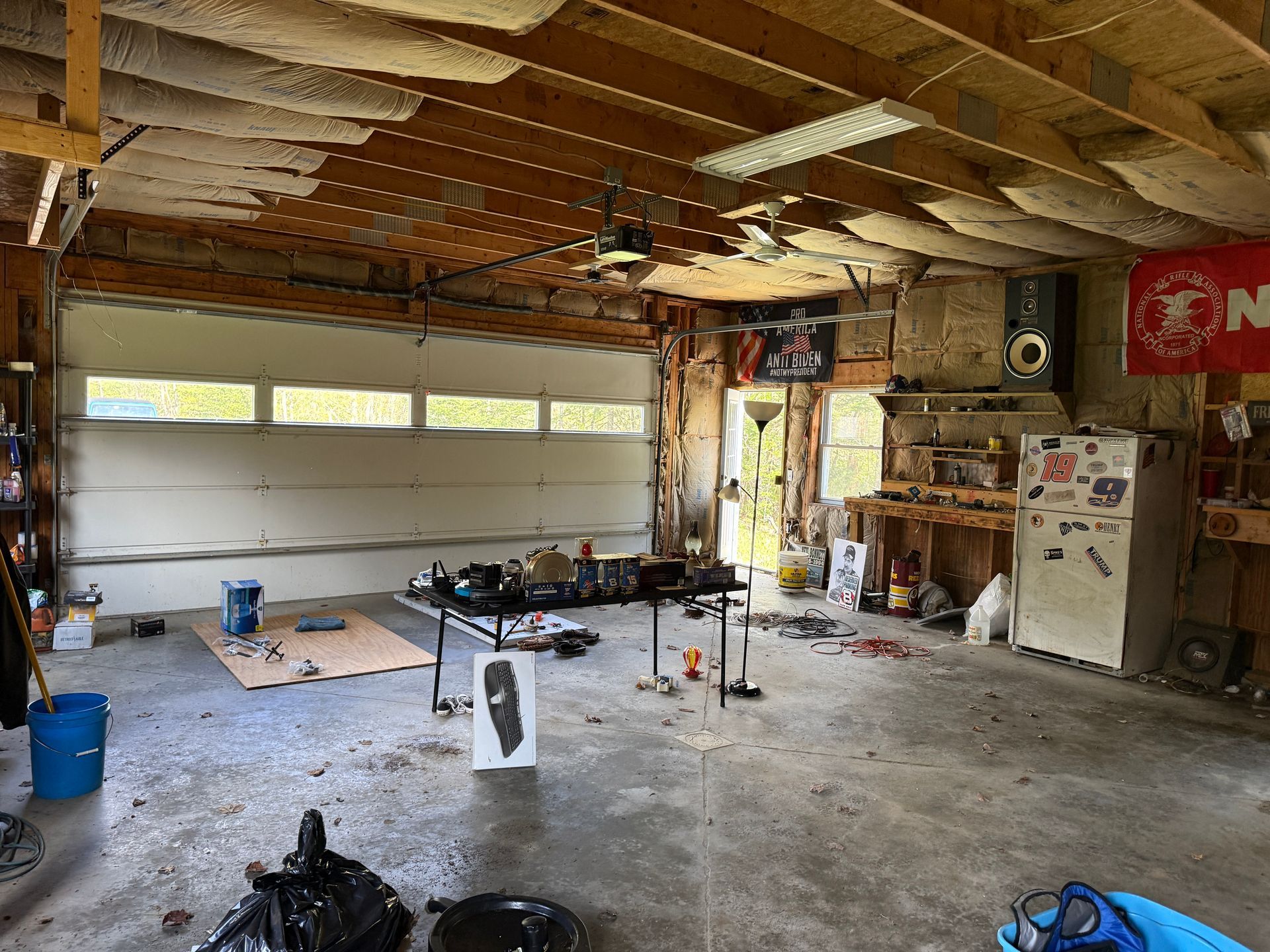 Garage with exposed ceiling, concrete floor, tools, shelves, and a refrigerator near the open door