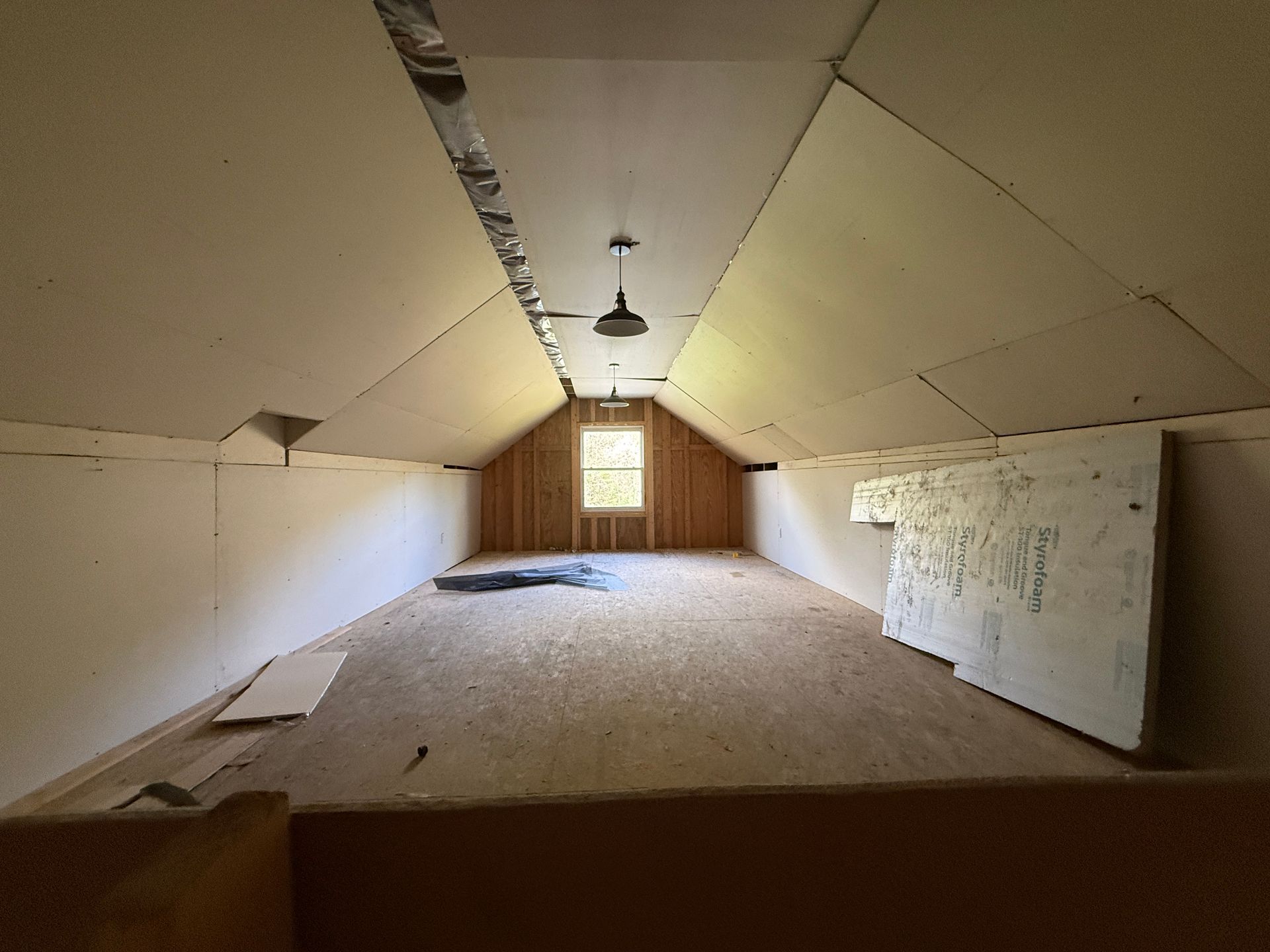 Attic-like empty room with sloped beige walls, carpeted floor, and a small window at the far end.