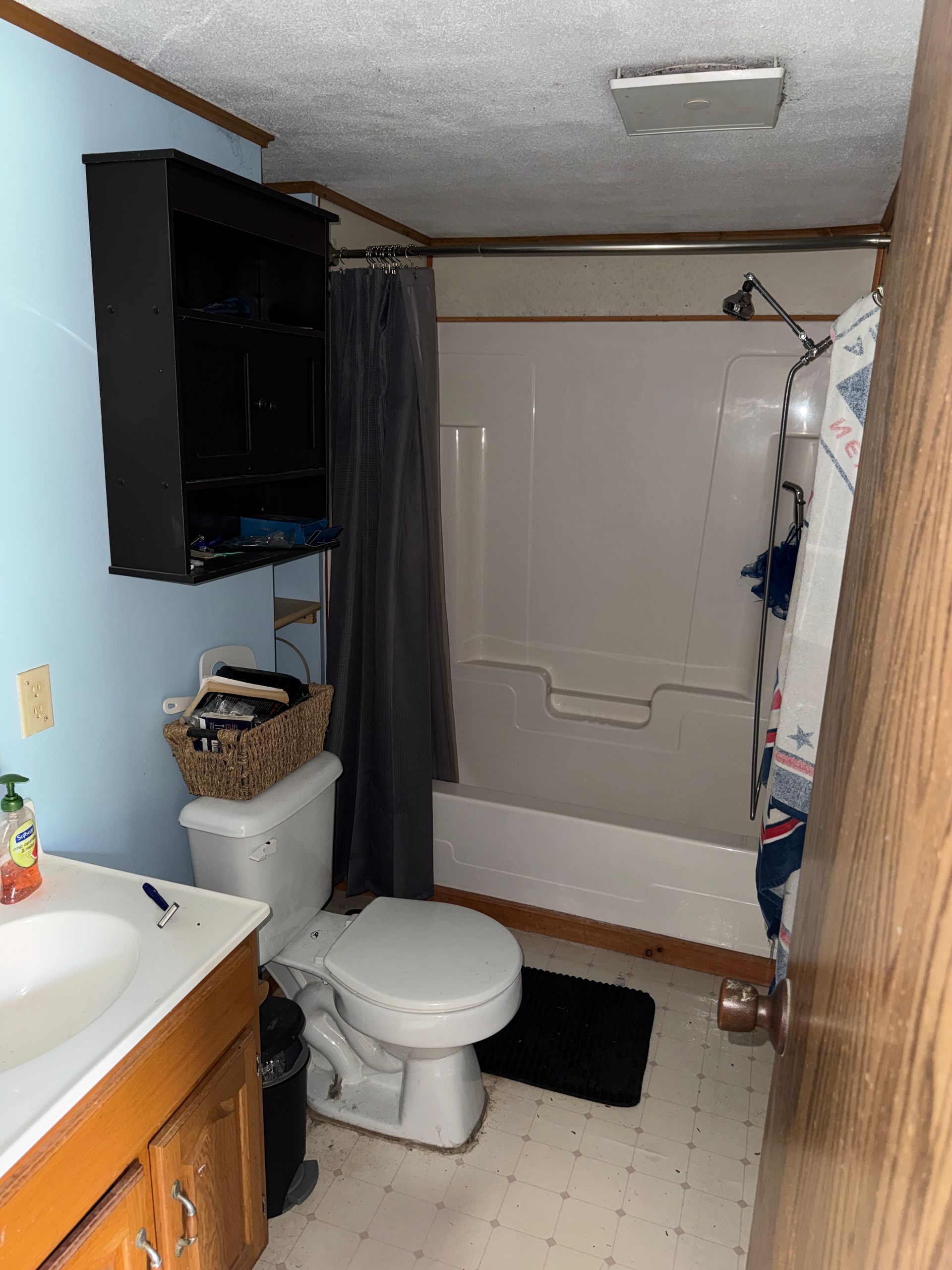 Small bathroom with toilet, shower curtain, sink, wooden cabinet, and black wall cabinet.