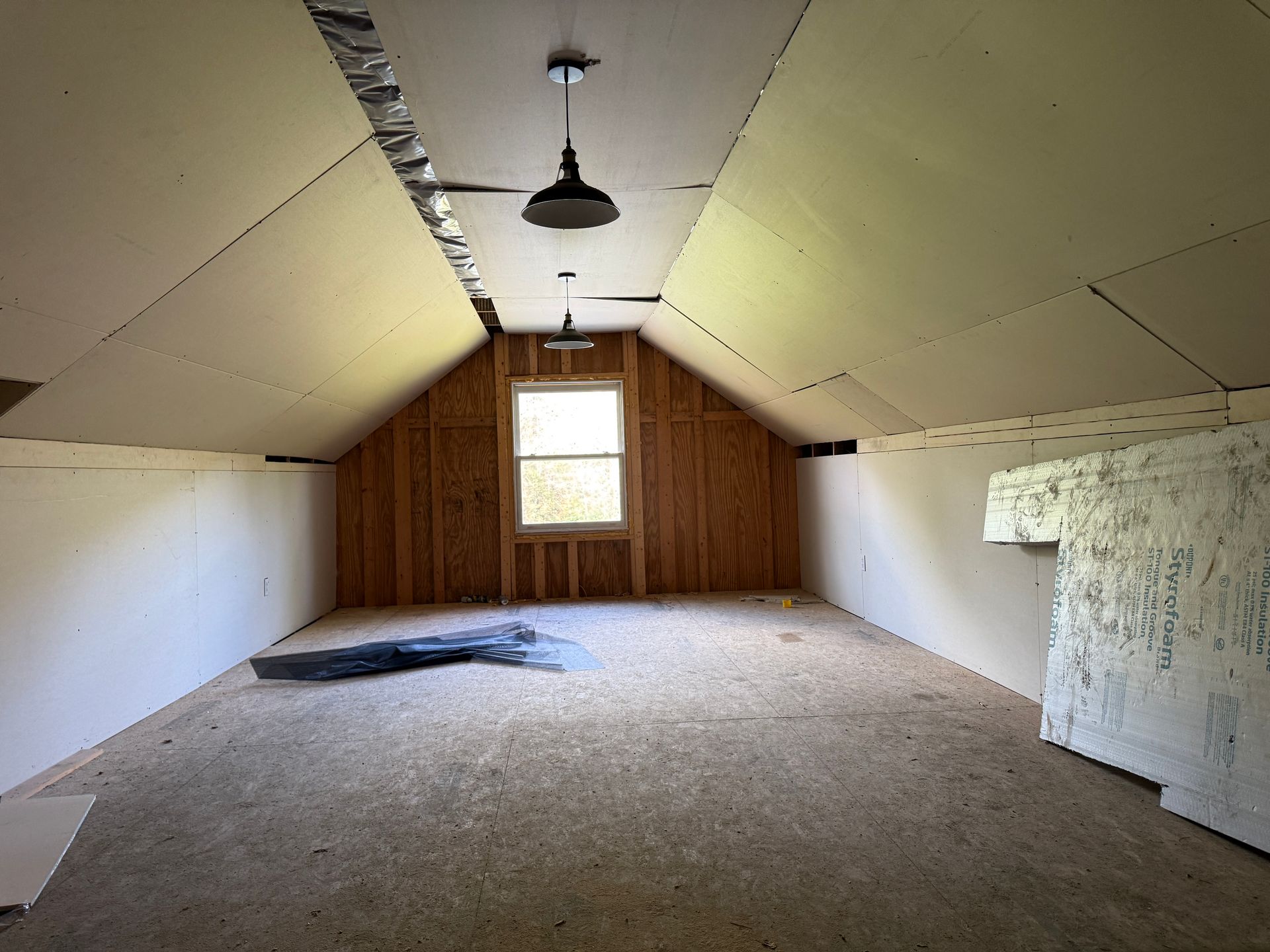 Empty attic room with sloped beige walls, wood-paneled far wall, and a single hanging light fixture