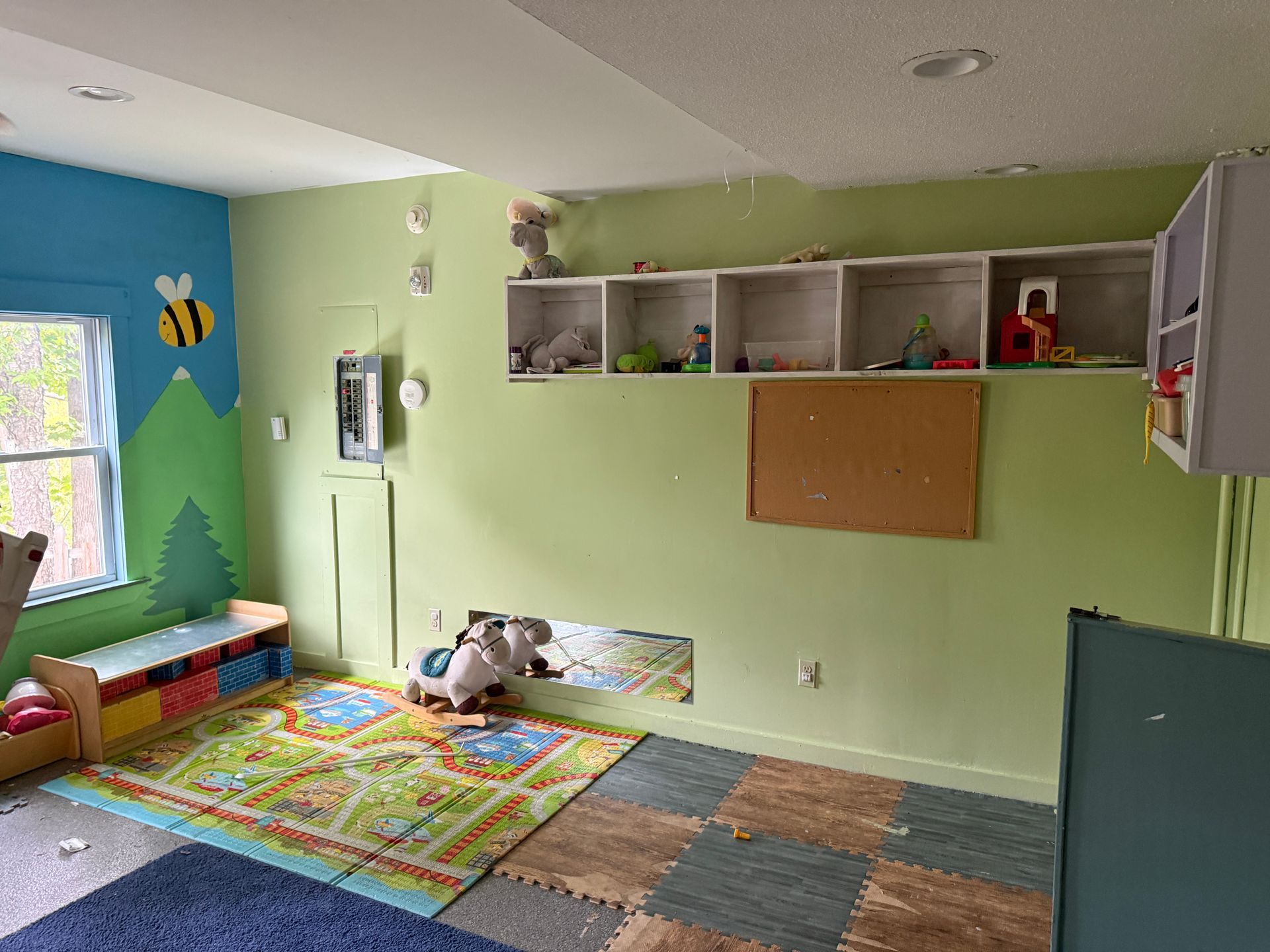 Colorful playroom with green walls, rugs, toys, shelves, and a window seat by the window.