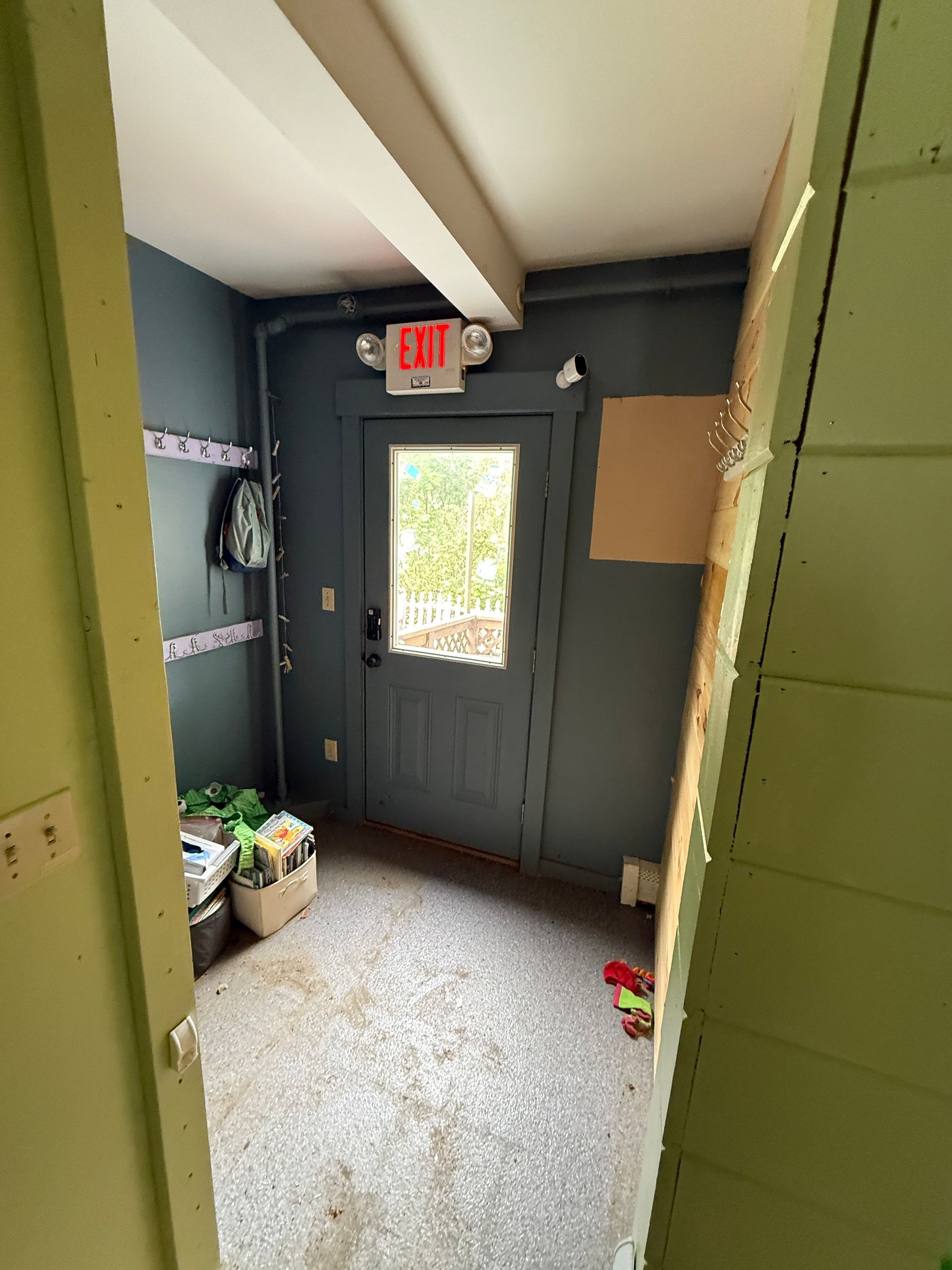 Narrow utility entryway with dark door, exit sign, speckled floor, and cleaning supplies on the left.