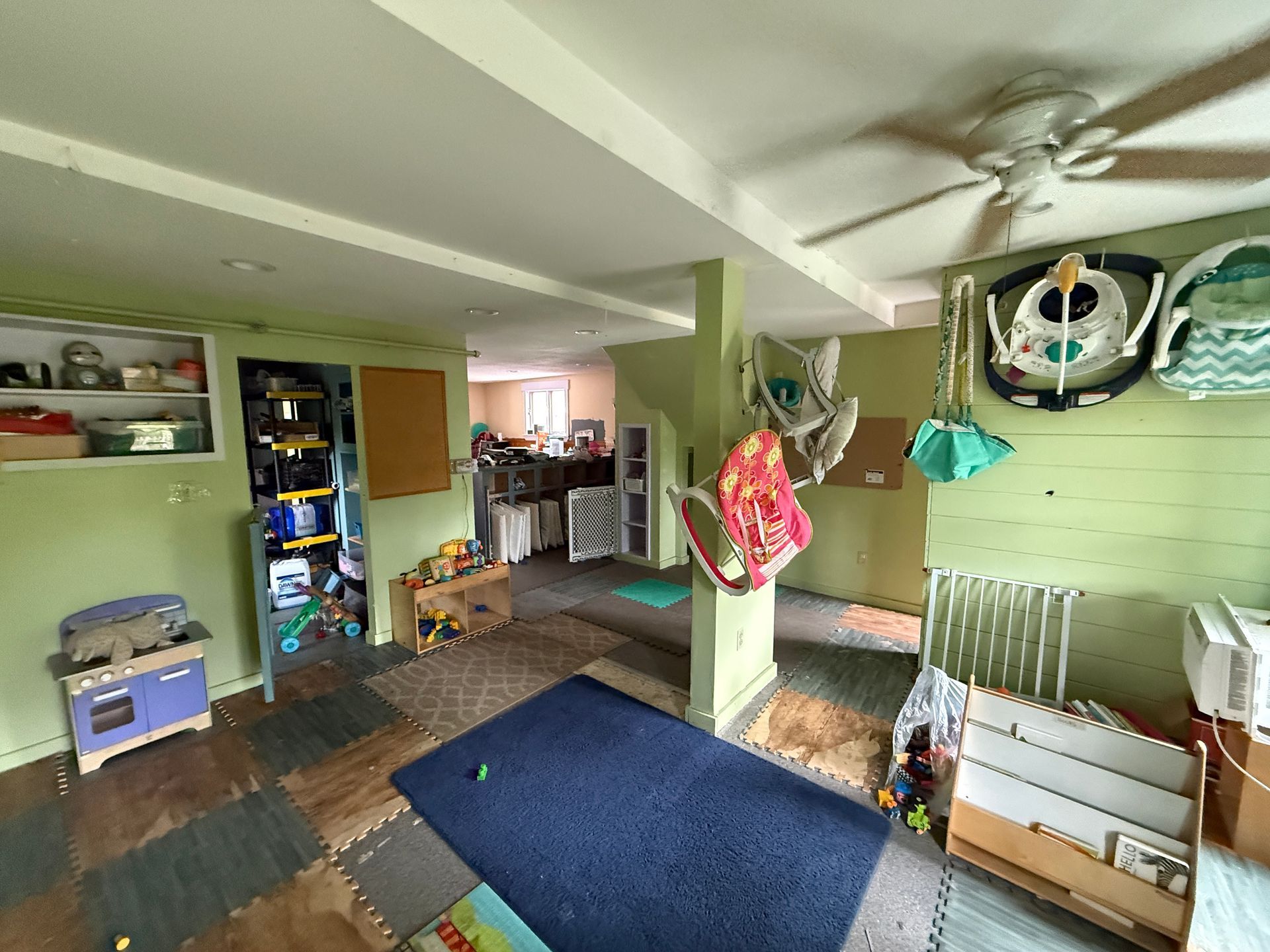 Green room with a blue rug, hanging toys, shelving, and an open doorway to a kitchen area