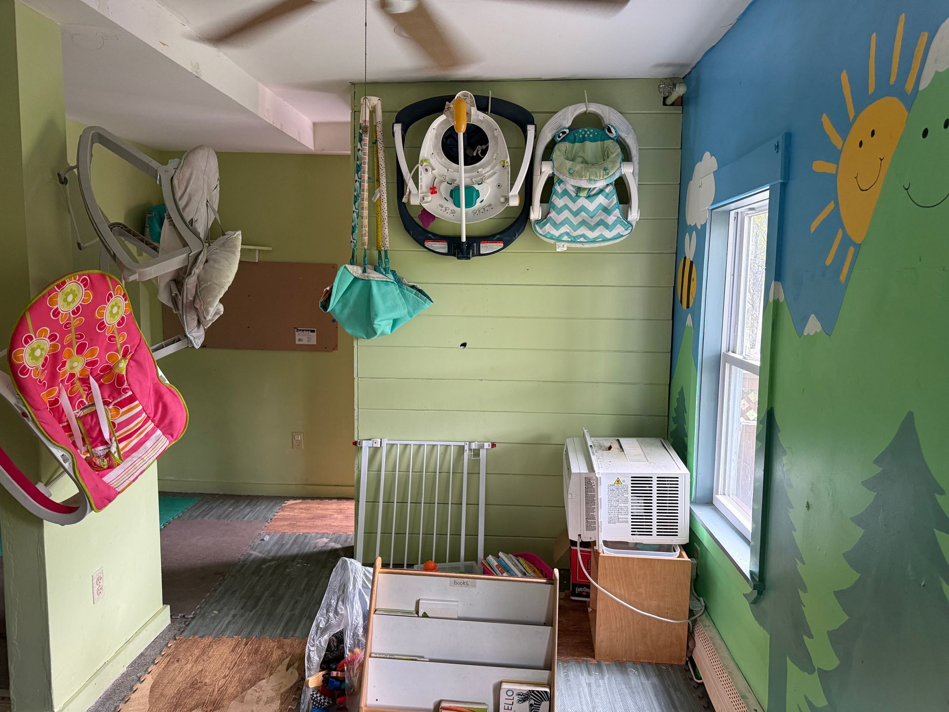 Colorful daycare room with hanging backpacks, crib, window, and sun mural on the wall