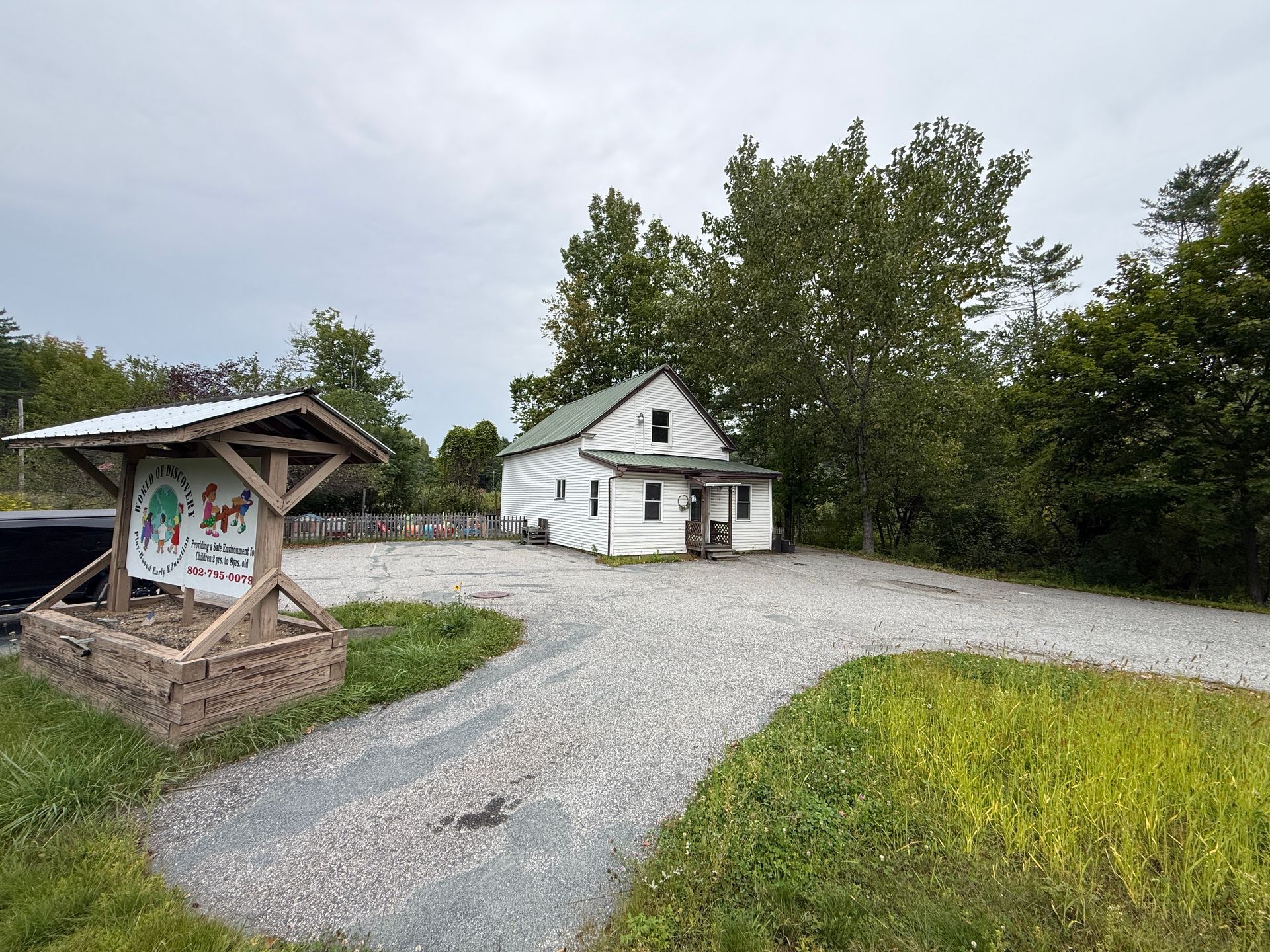 Rural gravel lot with a small white cabin and wooden sign under trees on an overcast day