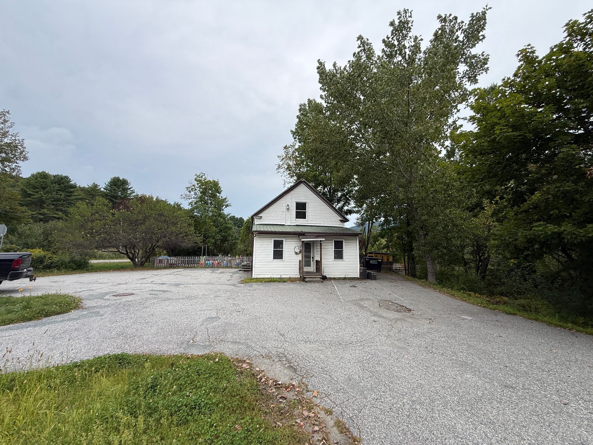 Small white house with a front porch, surrounded by gravel driveway and trees under a cloudy sky