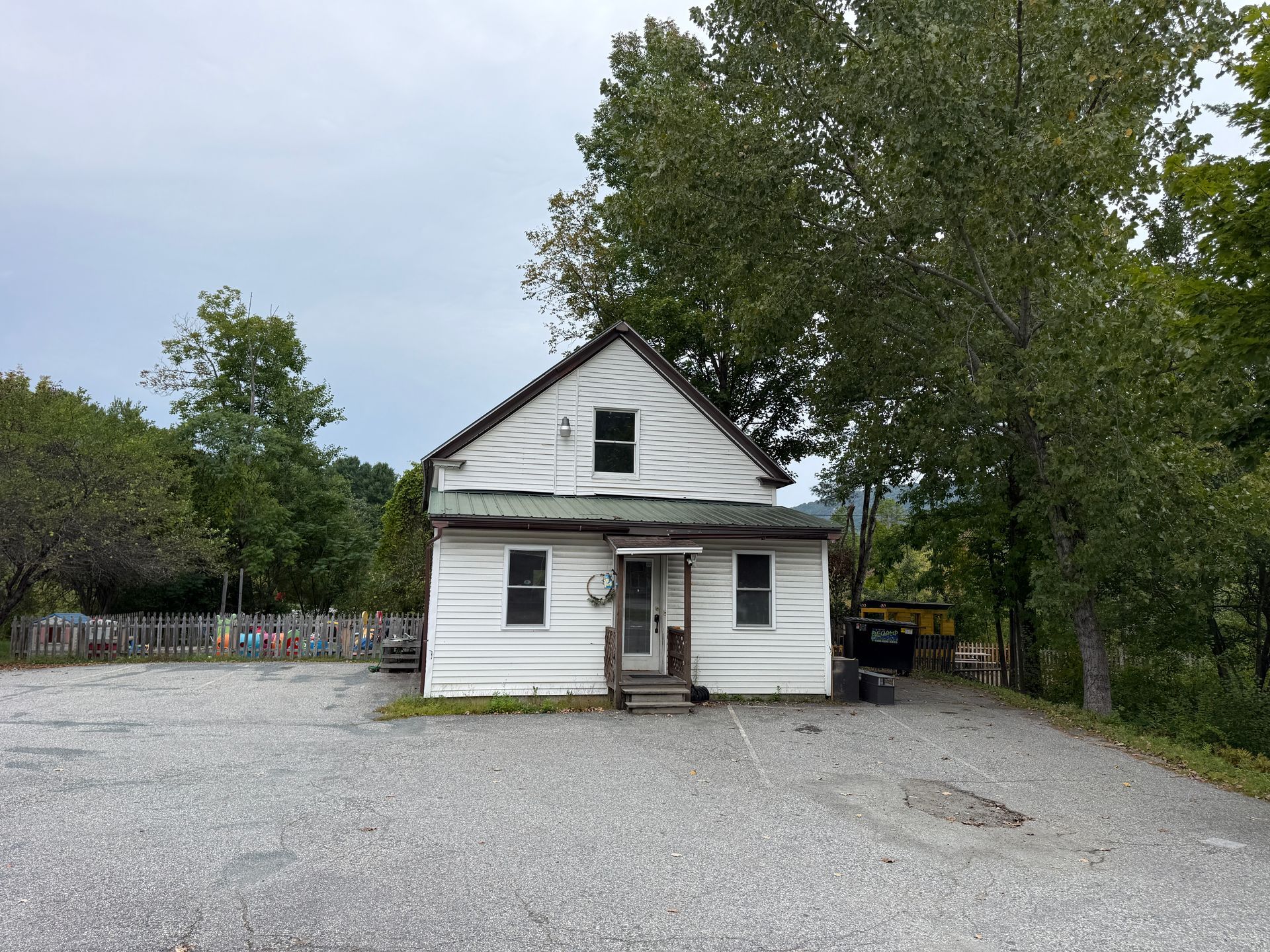 Small white house with a steep roof on a gravel lot, surrounded by trees under an overcast sky