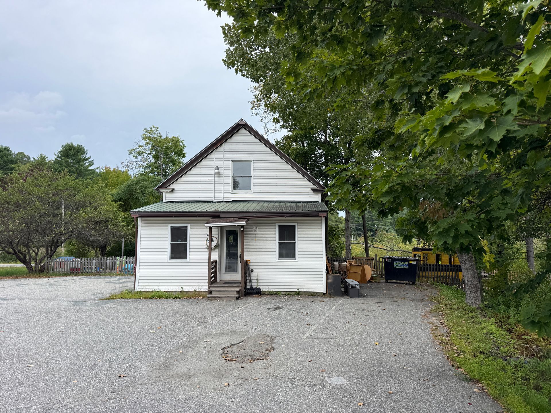 Small white house with a gravel driveway, trees, and a cloudy sky