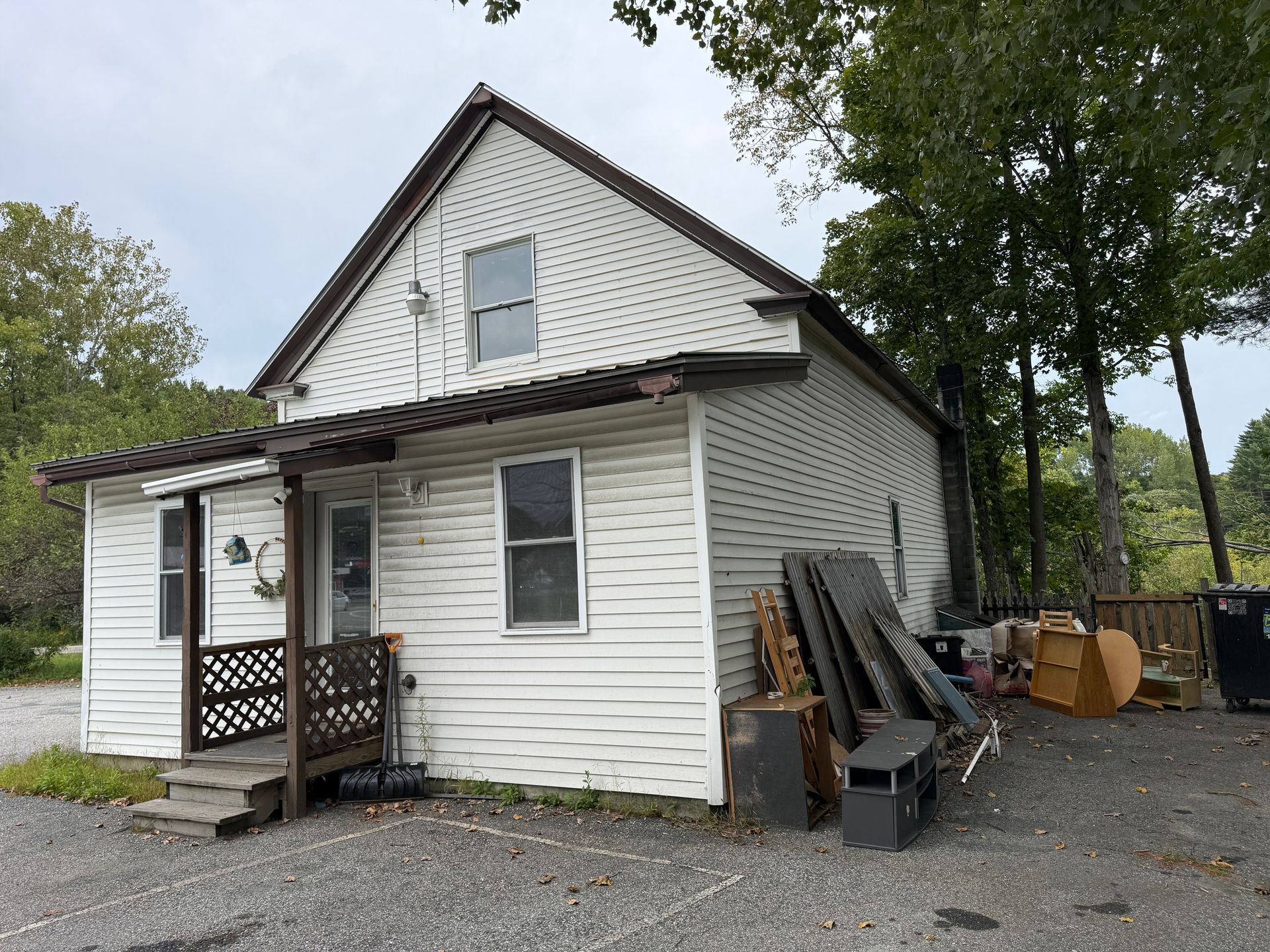 Small white house with porch, steep roof, and stacked items beside it on a gravel driveway.