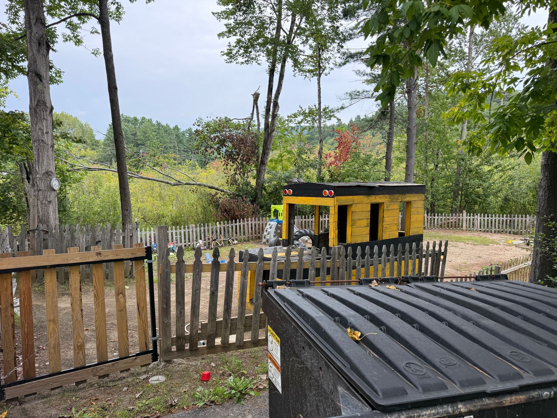 Wooded yard with a wooden fence and a yellow utility trailer