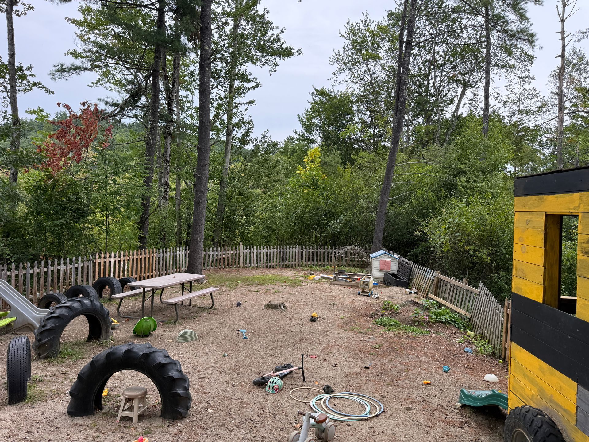 Outdoor play yard with tire obstacles, picnic table, wooden fence, and trees