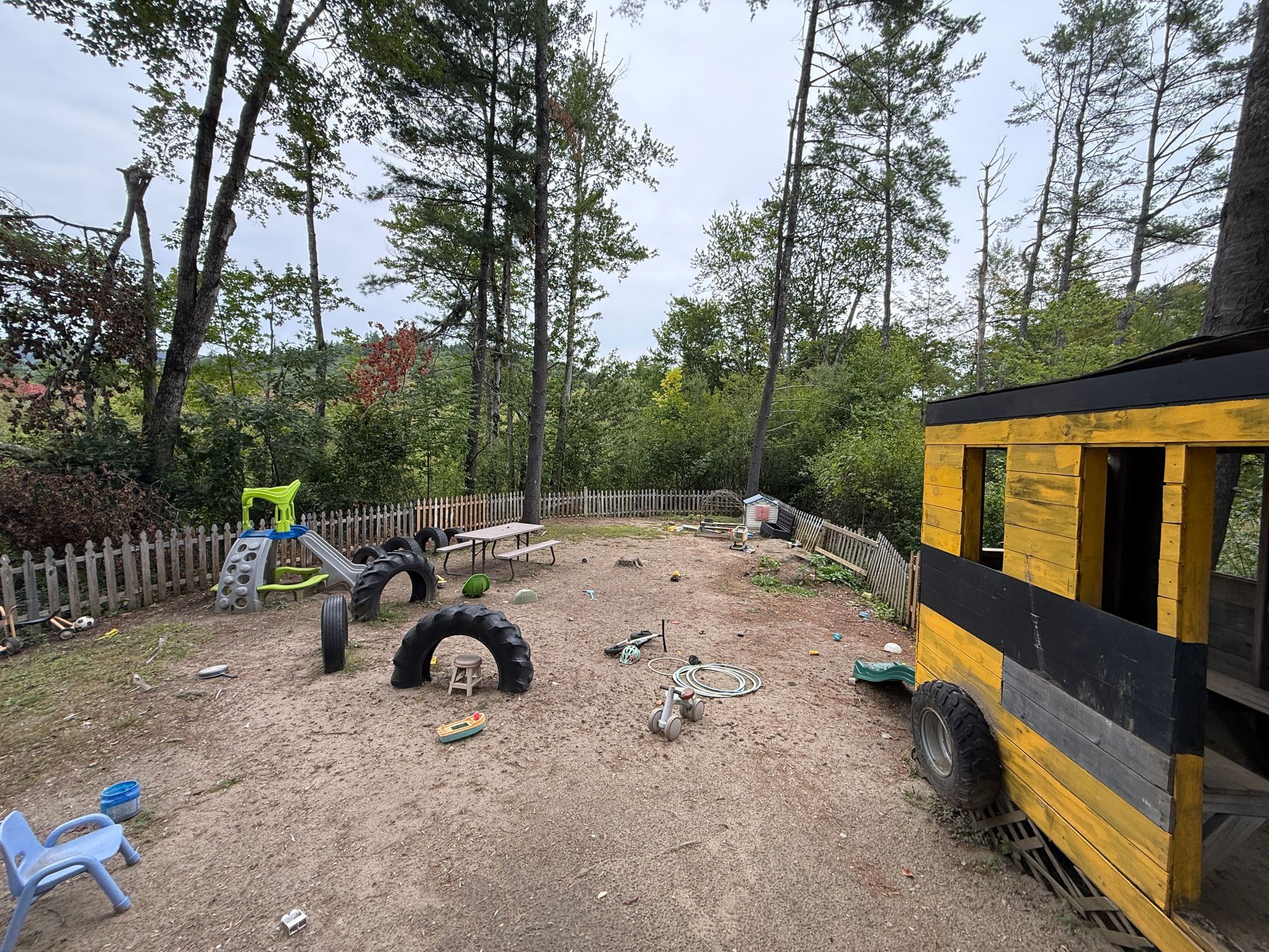 Backyard play area with tires, a blue chair, wooden fence, and a yellow-and-black structure among trees