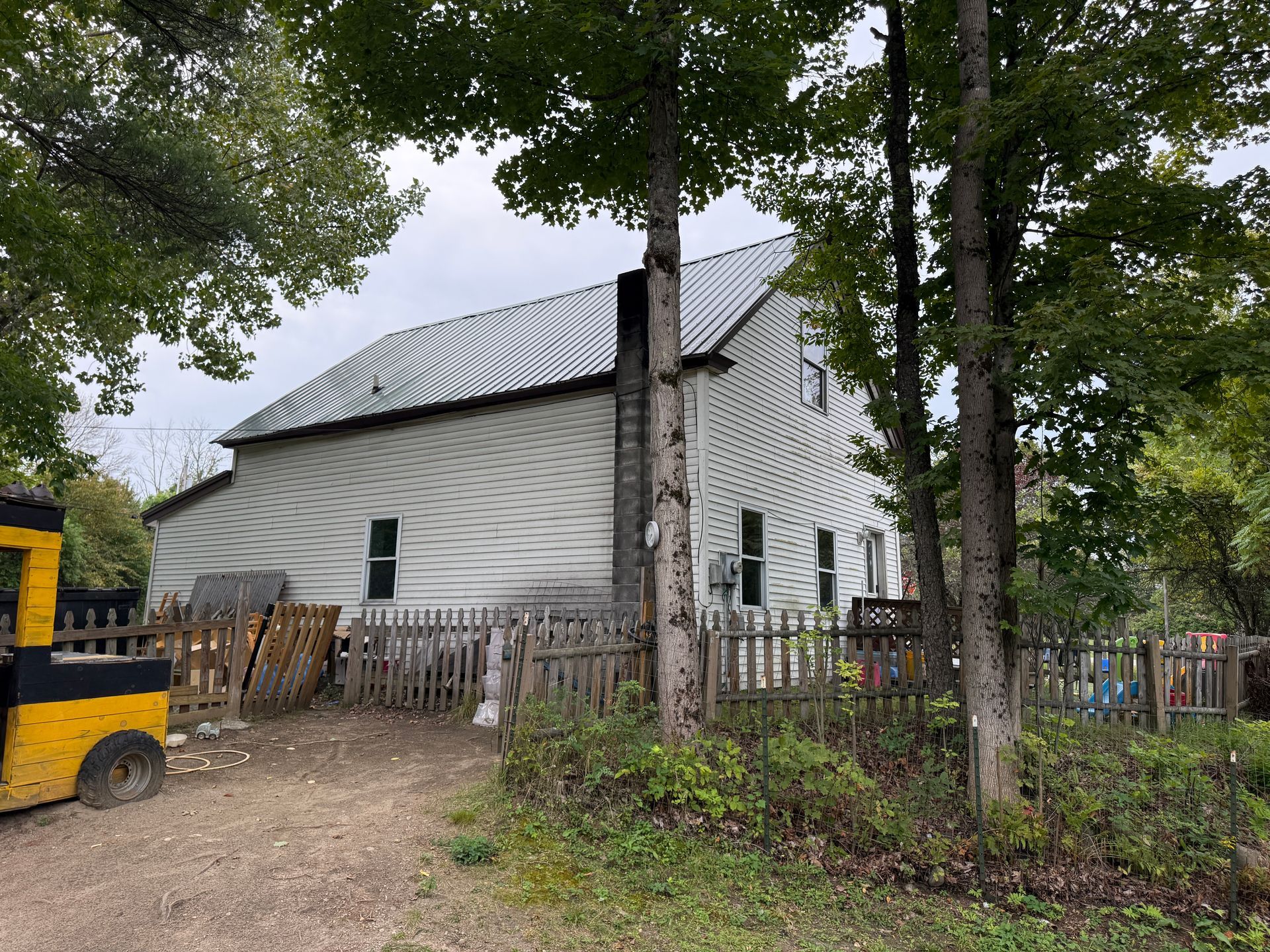 White house with a dark metal roof behind trees and a wooden fence, with a yellow vehicle at left.
