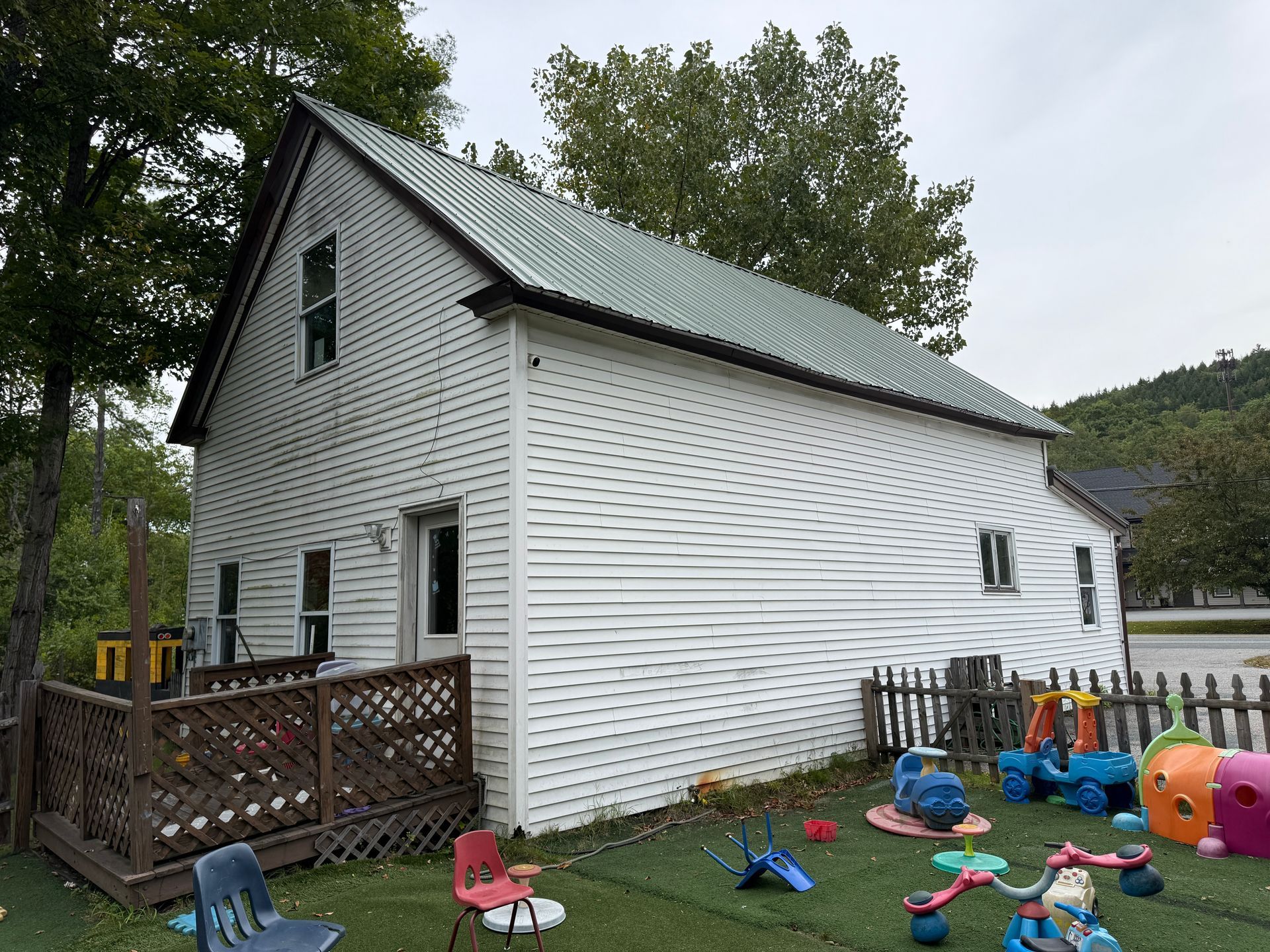 White two-story house with green roof and a fenced backyard, scattered with colorful children's toys and a small deck.