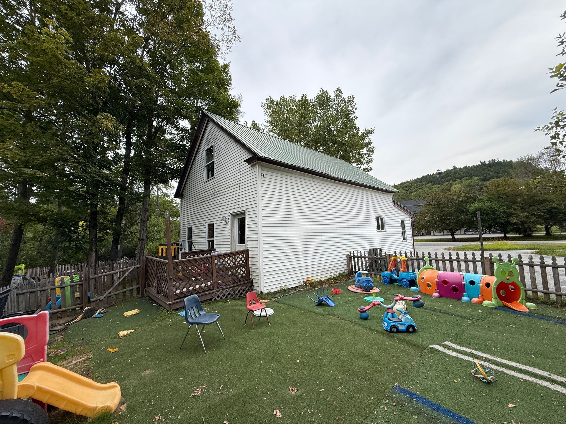 Small white house with siding beside a playground of colorful toys and a slide on grass.