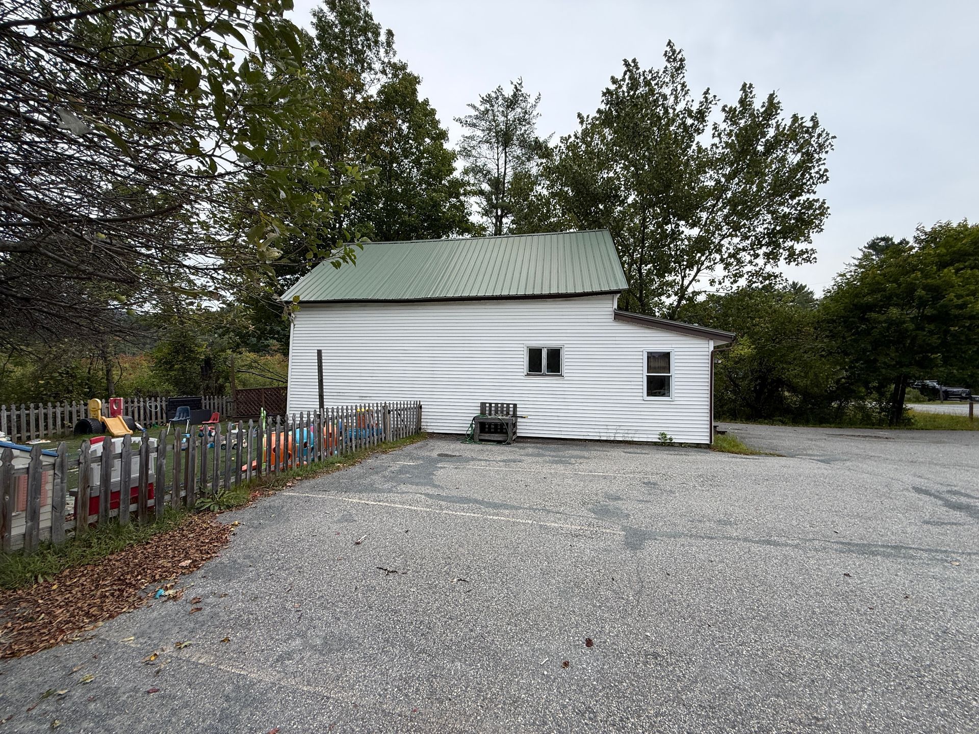 Small white building with green roof beside a gravel lot and chain-link fence, surrounded by trees
