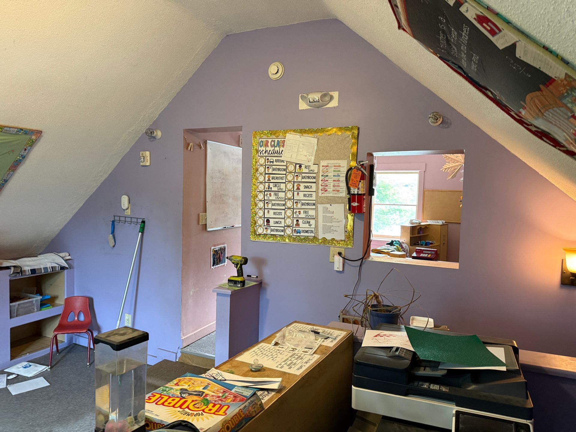 Cozy attic office with purple walls, desk, bulletin board, lamp, and sloped ceiling windows.