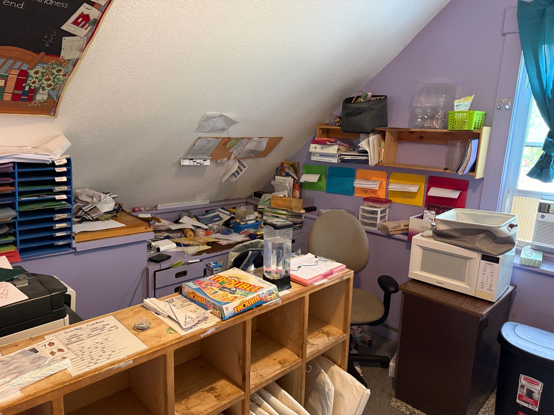attic office with shelves, stacks of papers, a desk, and a window with blue curtains.