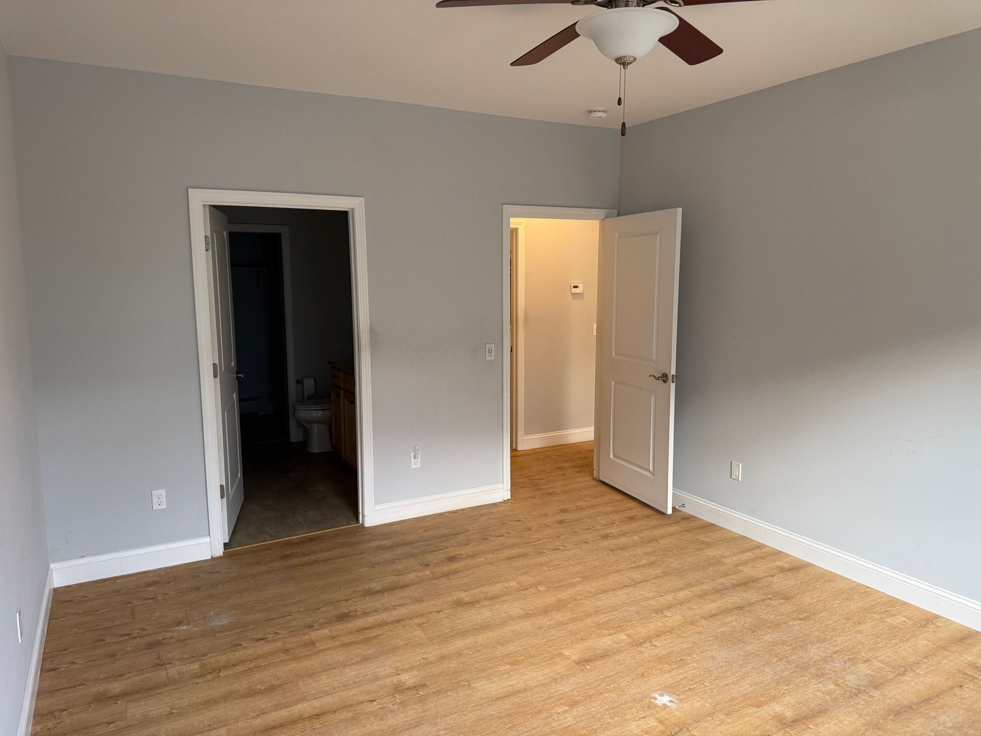 Empty bedroom with gray walls, wood floor, open closet, and open door to a lit hallway.