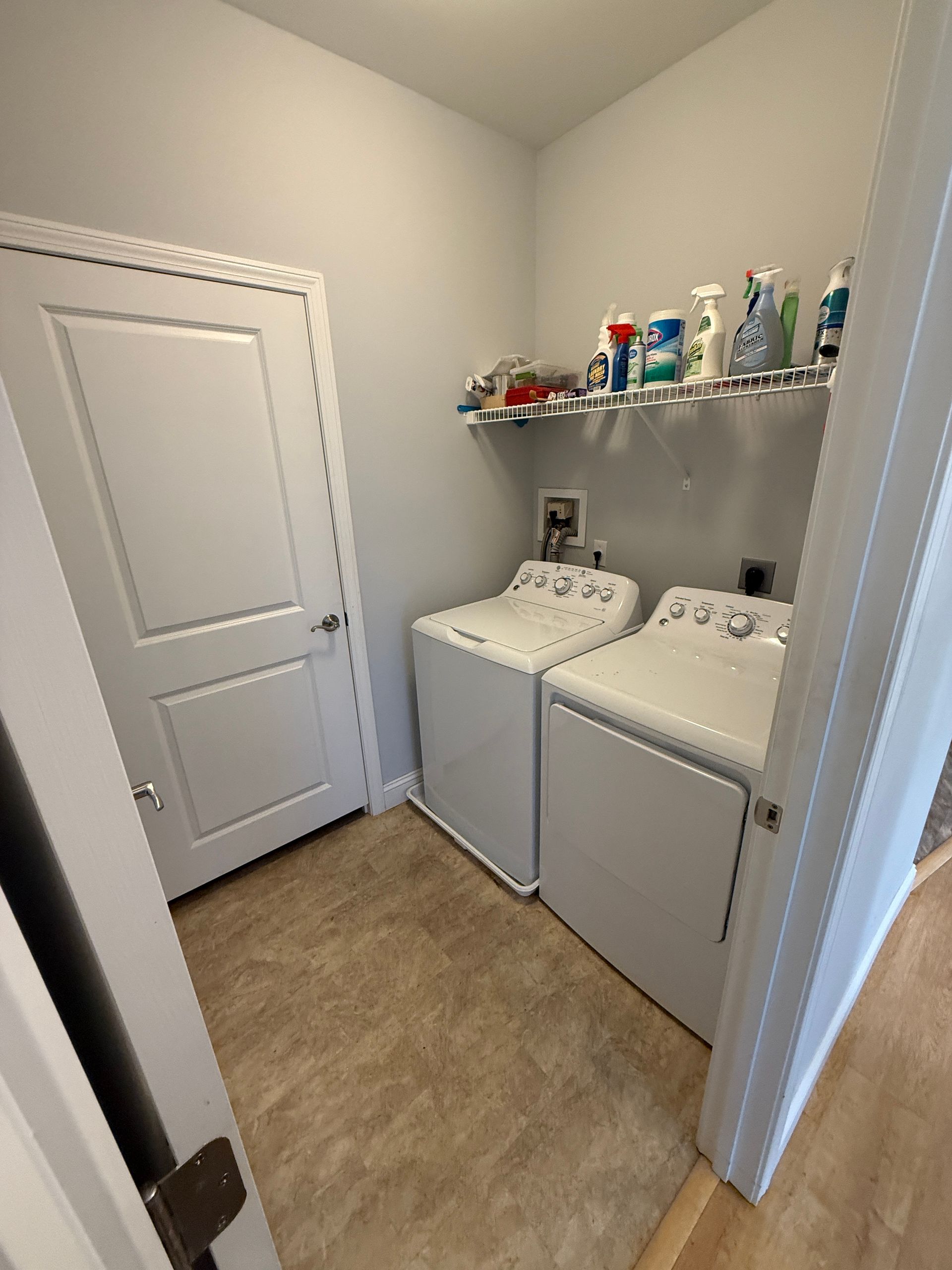 Small laundry room with two white washers, detergent shelf, and a closed door.