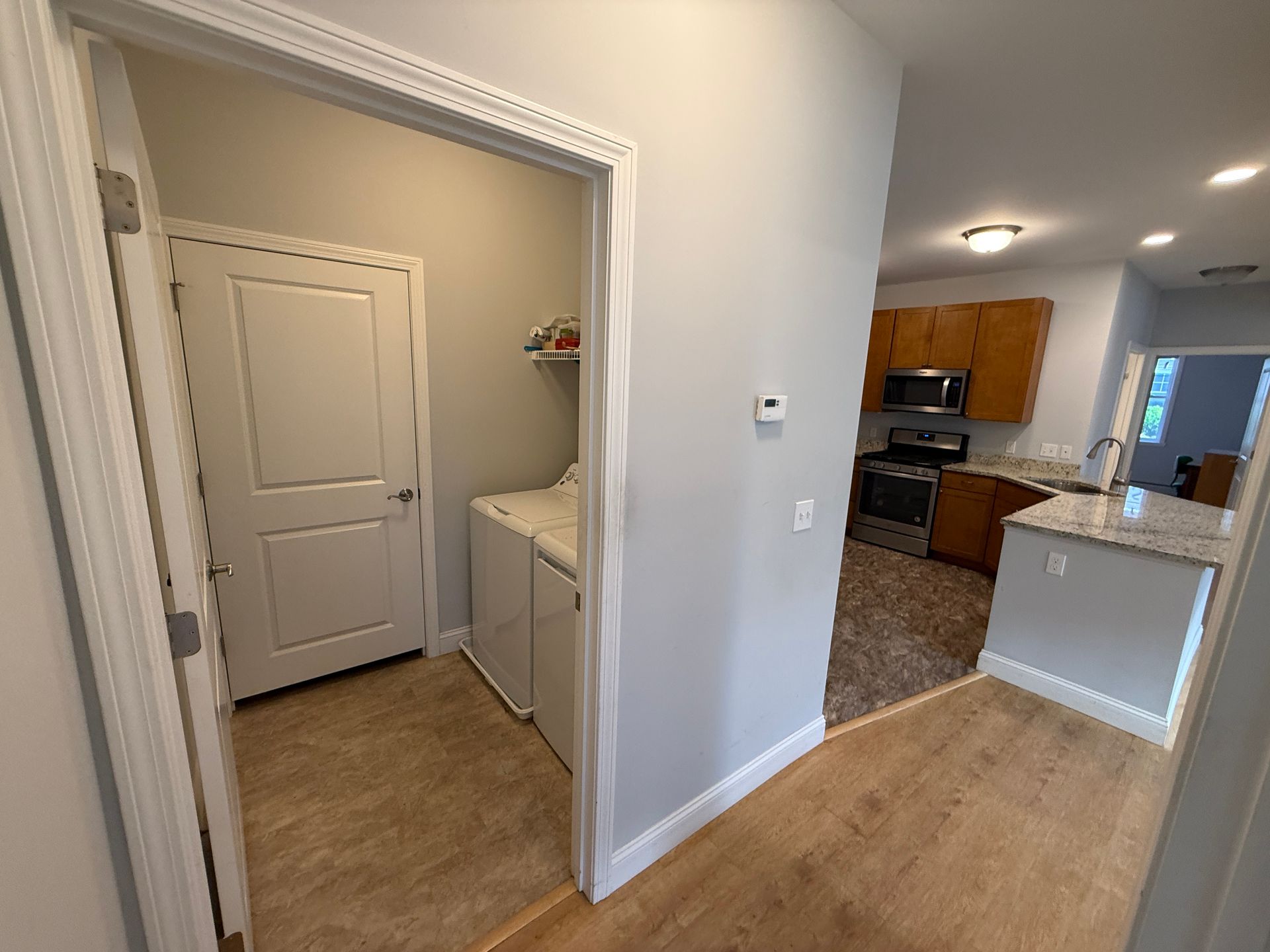 Hallway view into laundry room and kitchen with beige flooring and white walls