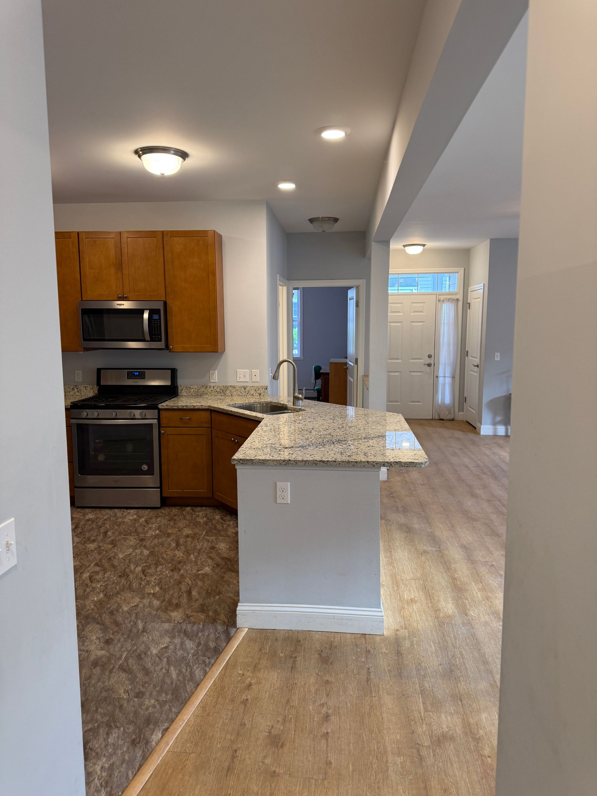 Empty kitchen and hallway in a home with wood cabinets, granite island, and light wood flooring