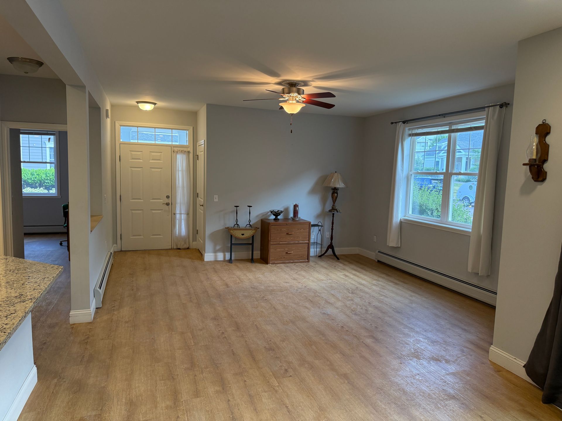 Empty living room with wood floors, front door, large windows, and a ceiling fan.