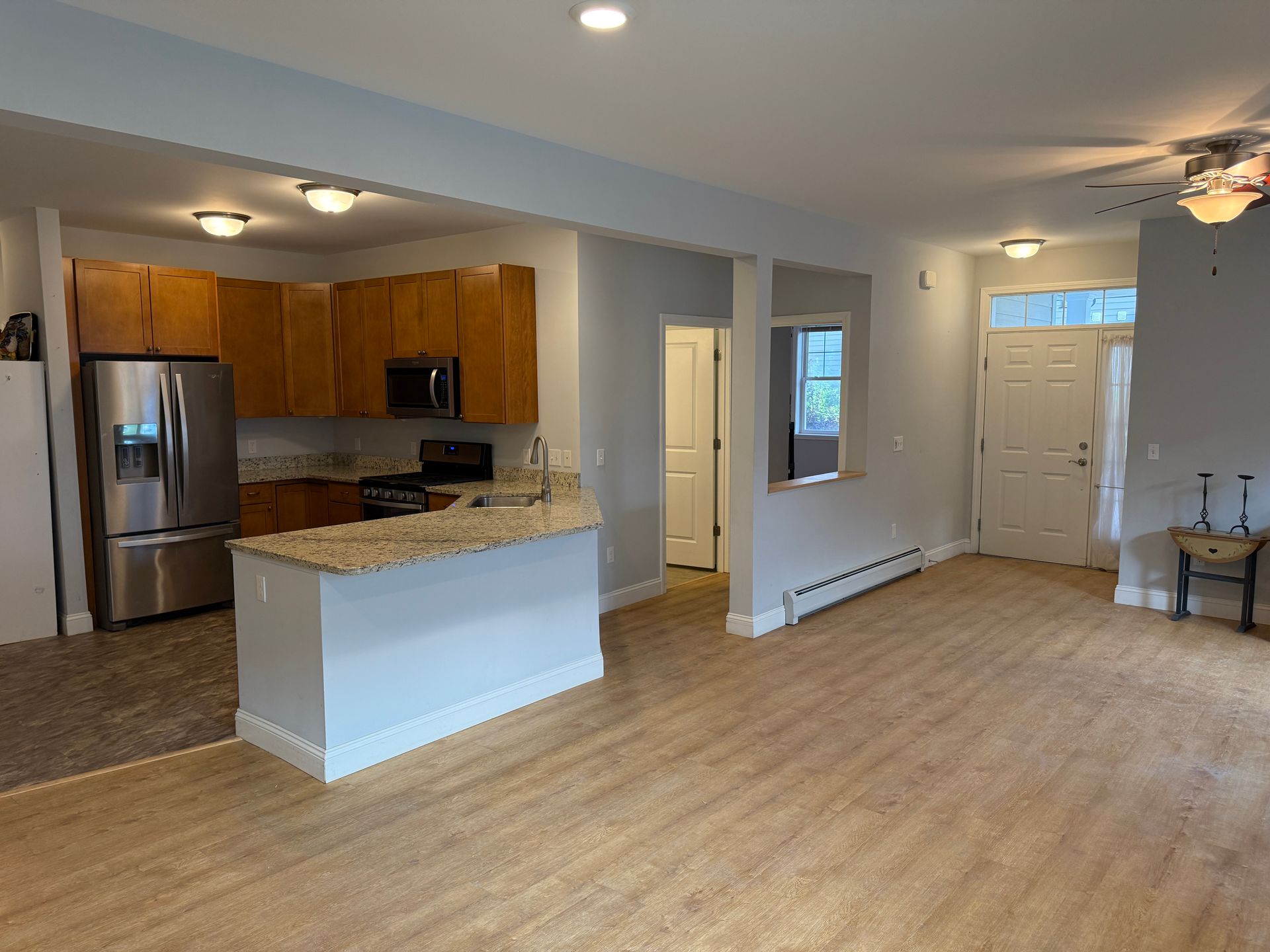 Empty open-plan kitchen and living area with wood cabinets, stainless fridge, and light wood flooring