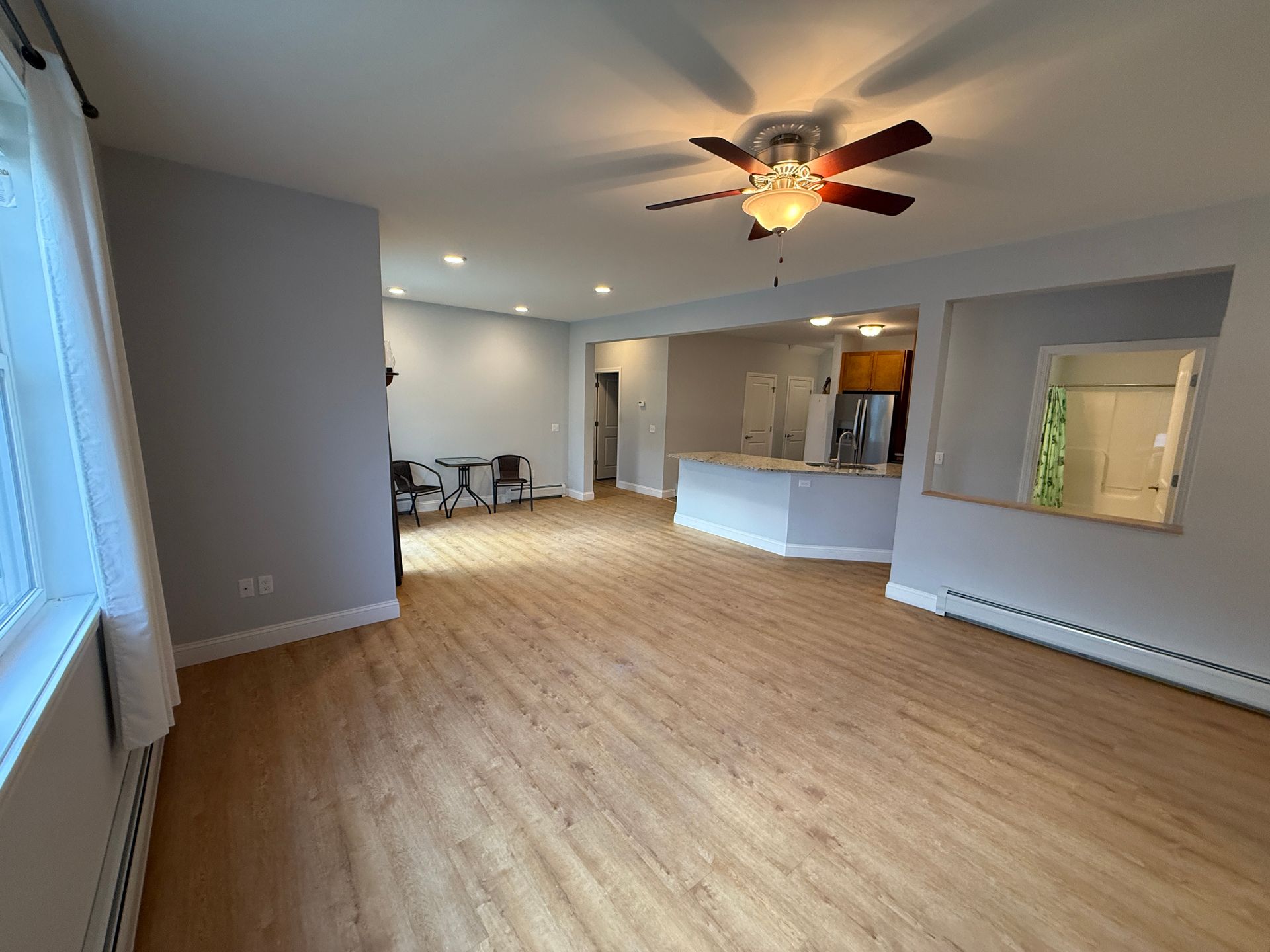 Empty open-plan living room with wood floors, gray walls, ceiling fan, and a kitchen passthrough.