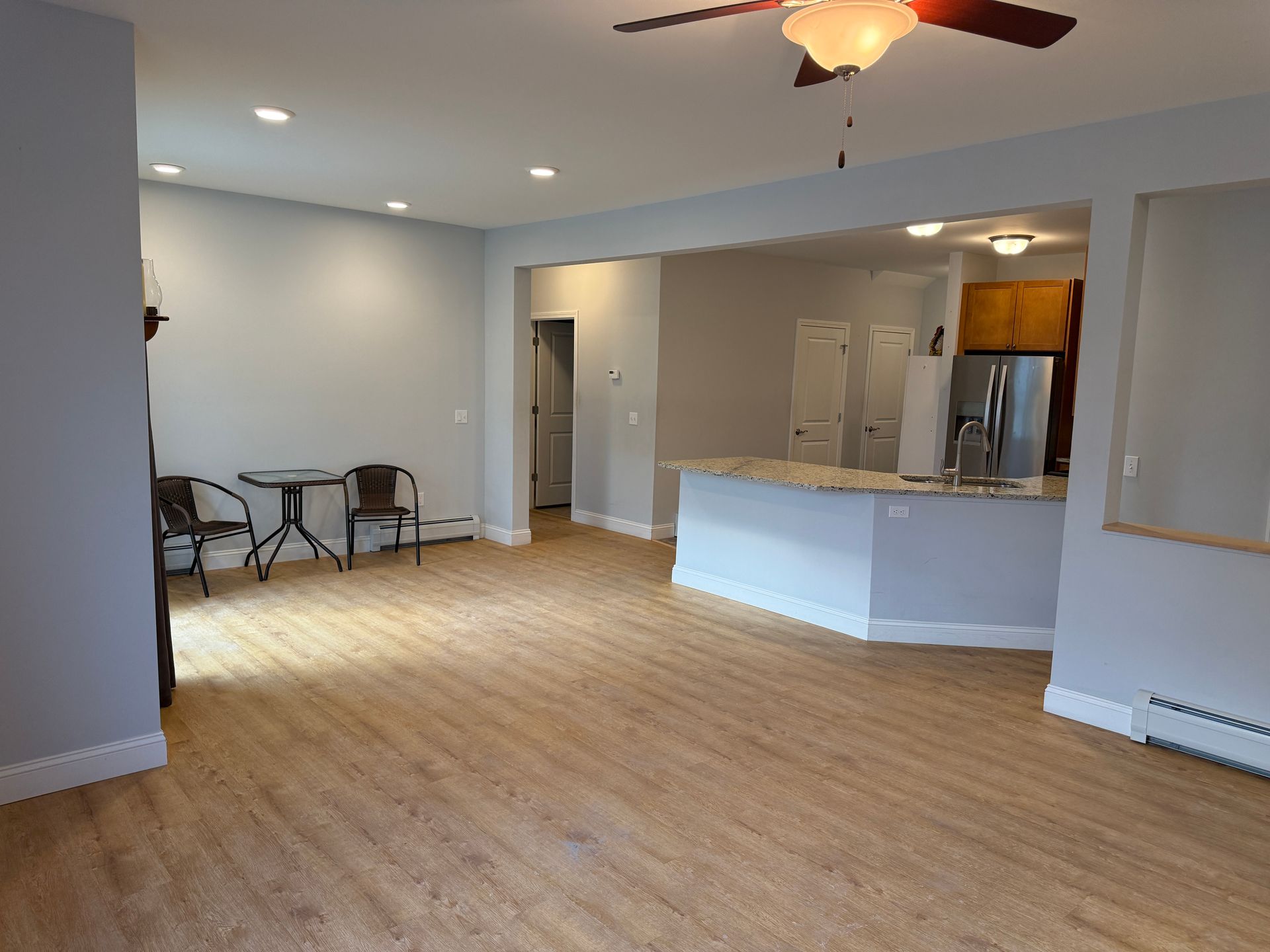 Empty open-plan living room with light wood floors, gray walls, ceiling fan, and kitchen area with island.