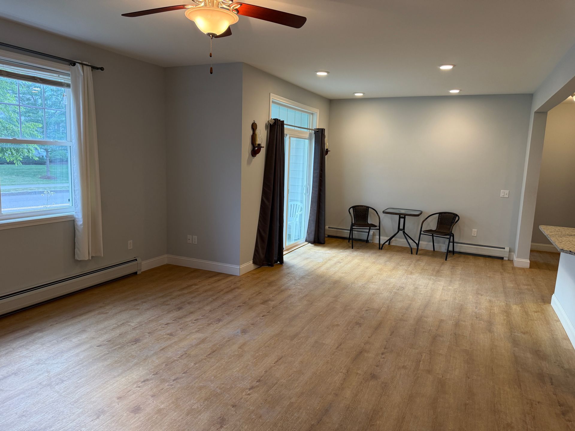 Empty beige living room with hardwood floors, ceiling fan, windows, and a small table with chairs.