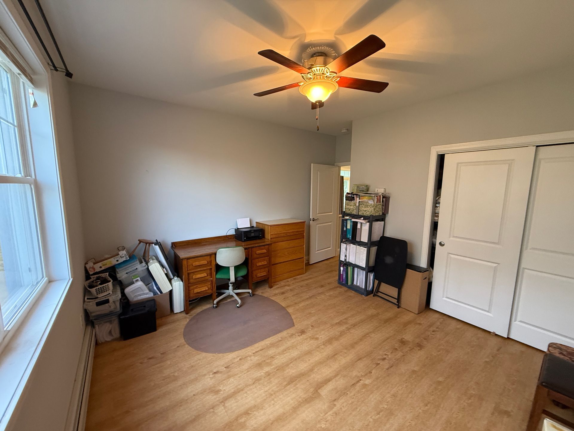 Small home office with desk, chair, storage bins, wood floor, ceiling fan, and closet doors.