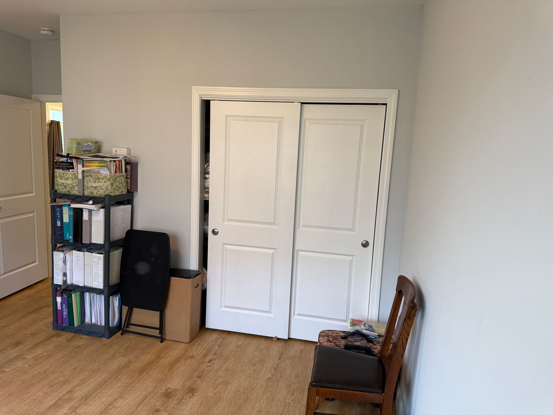 Empty room with white double doors, a bookshelf, folded chair, and wooden chair on a hardwood floor.