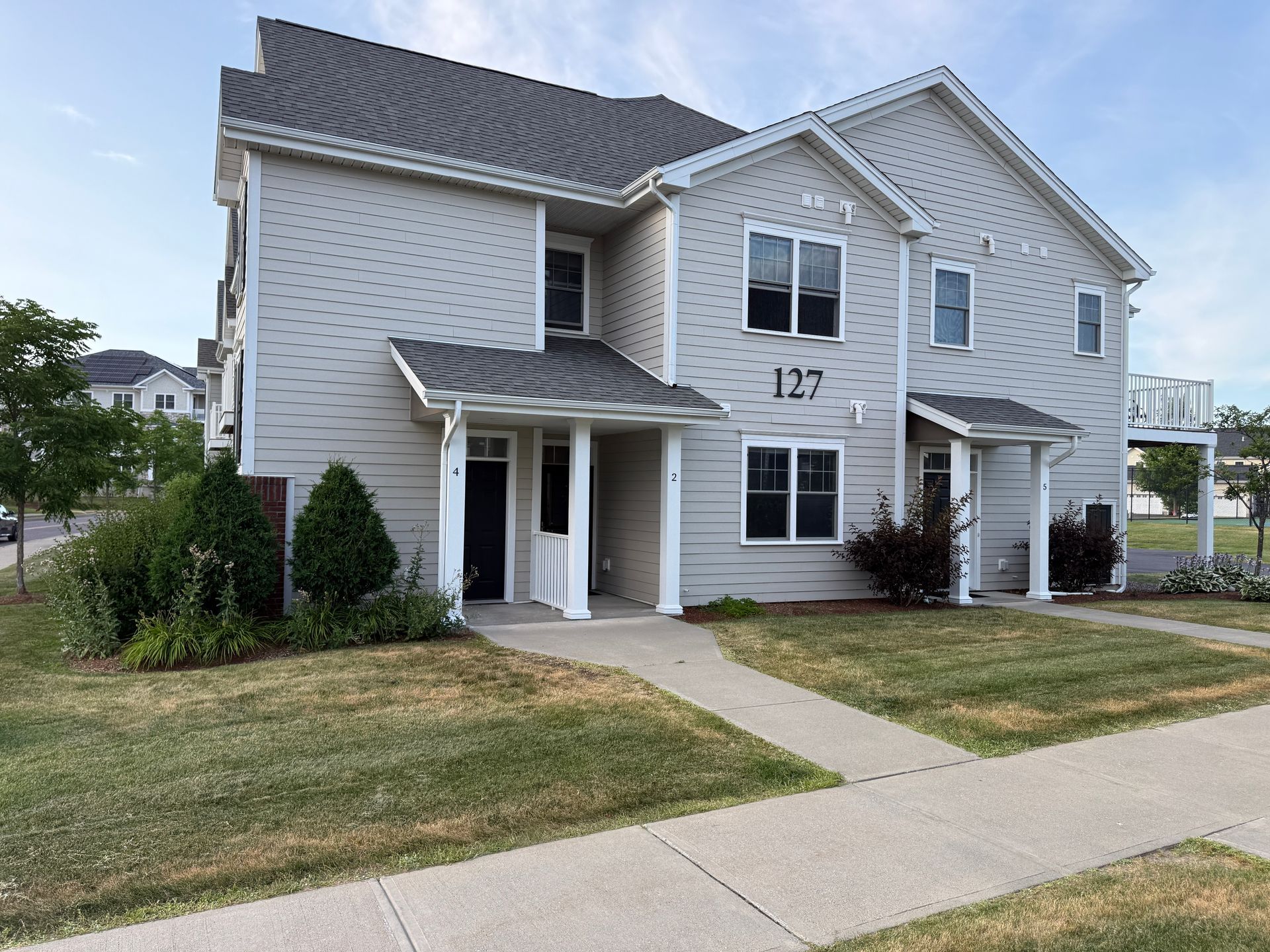 White two-story townhouse with gray roof, front porch, small lawn, and sidewalk in a suburban neighborhood