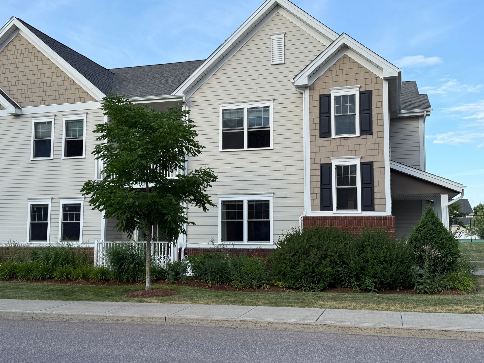 Suburban townhouse with beige siding, white trim, and dark brick accents beside a street and trees