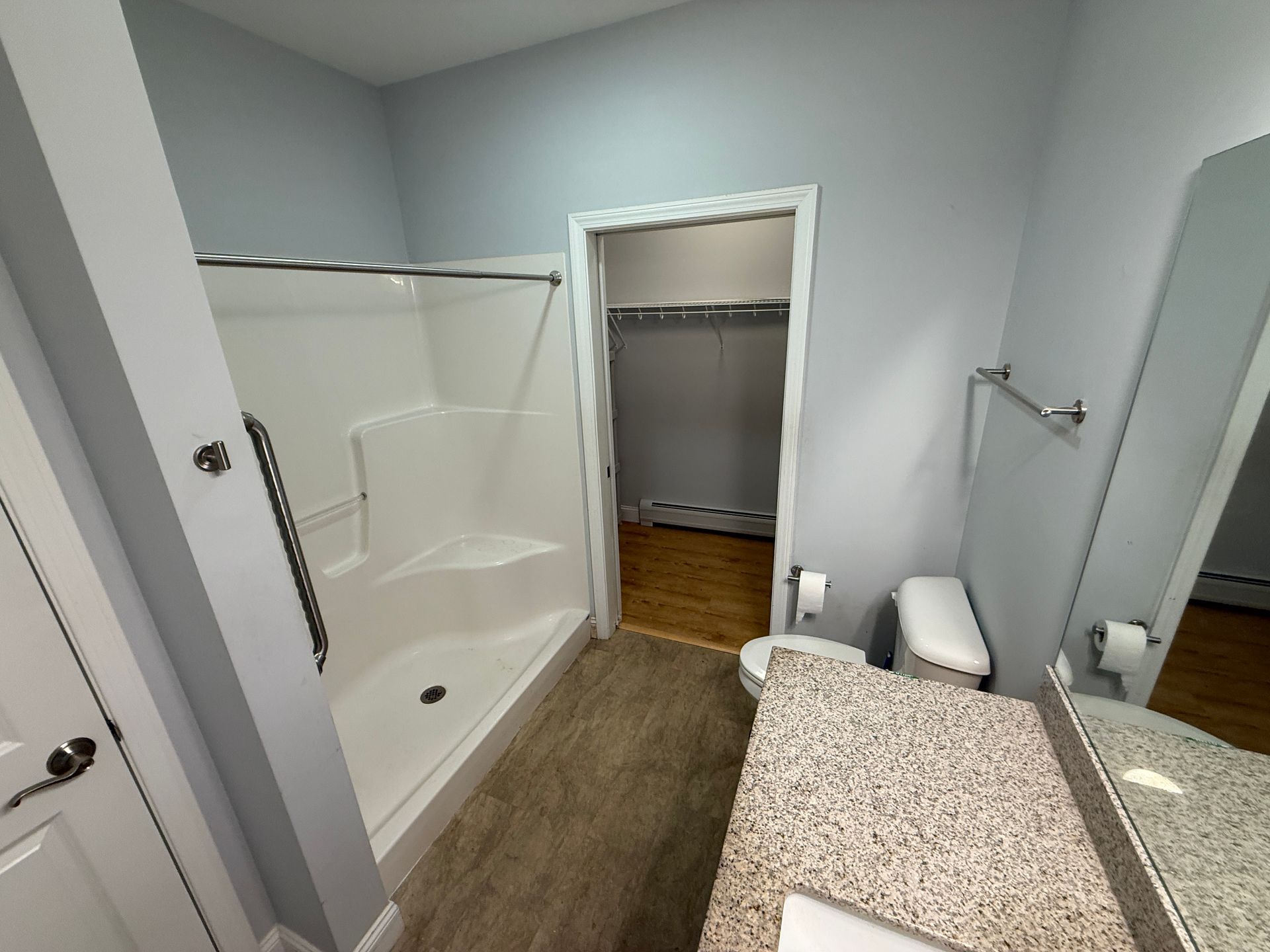 Bathroom with walk-in shower, vanity, toilet, and open closet door to a wood-floored room.
