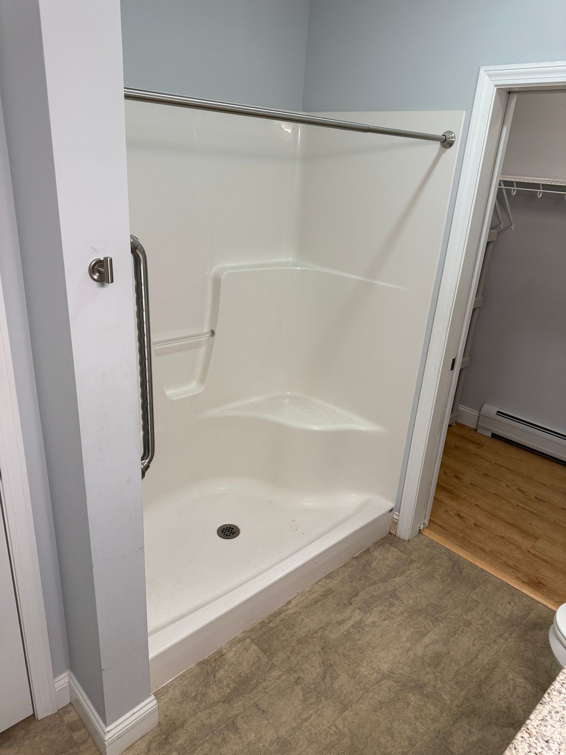 Empty shower stall with white walls, grab bars, and a curtain rod beside beige carpet and wood flooring