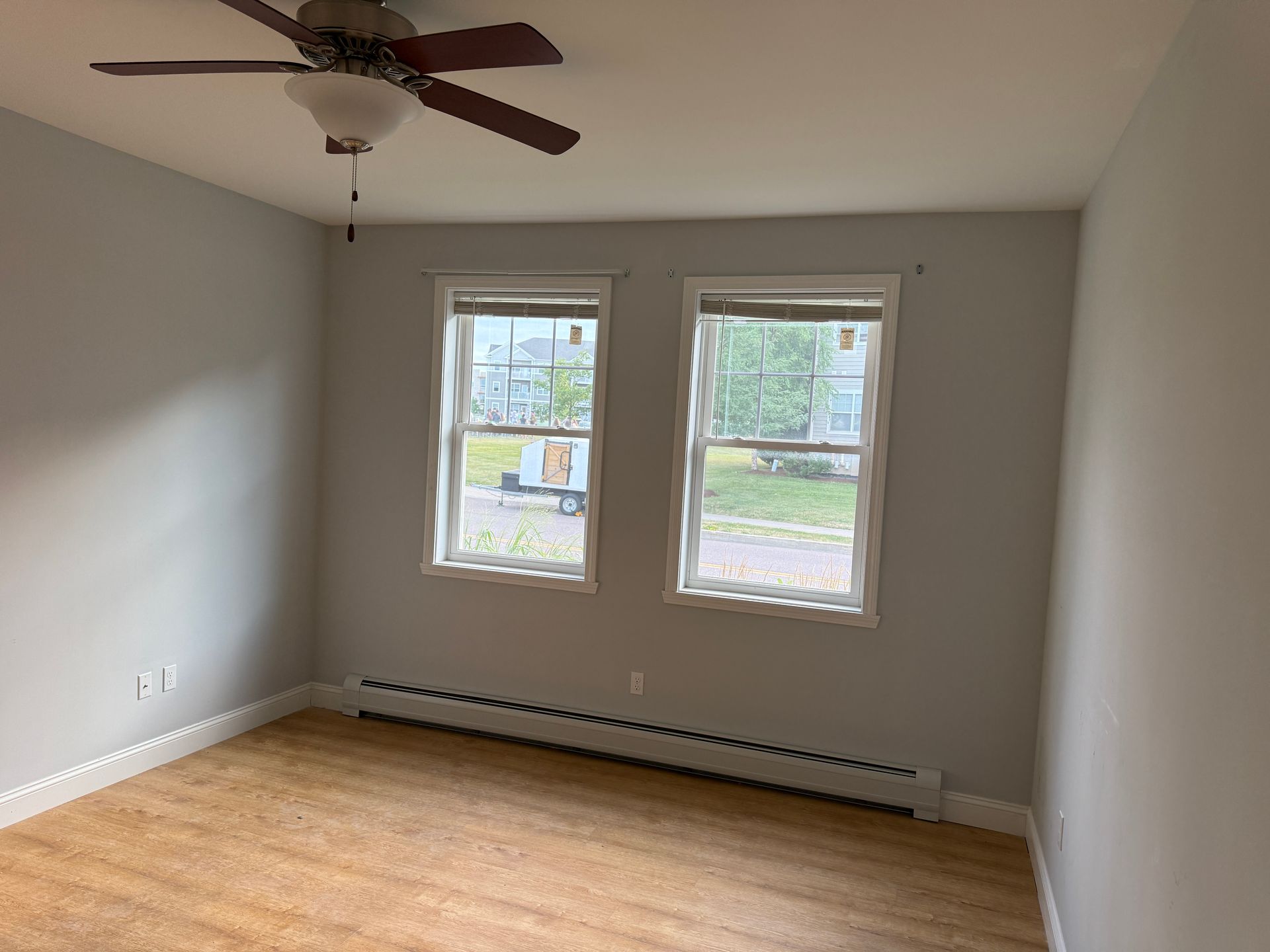 Empty room with light wood floors, gray walls, two windows, and a ceiling fan.