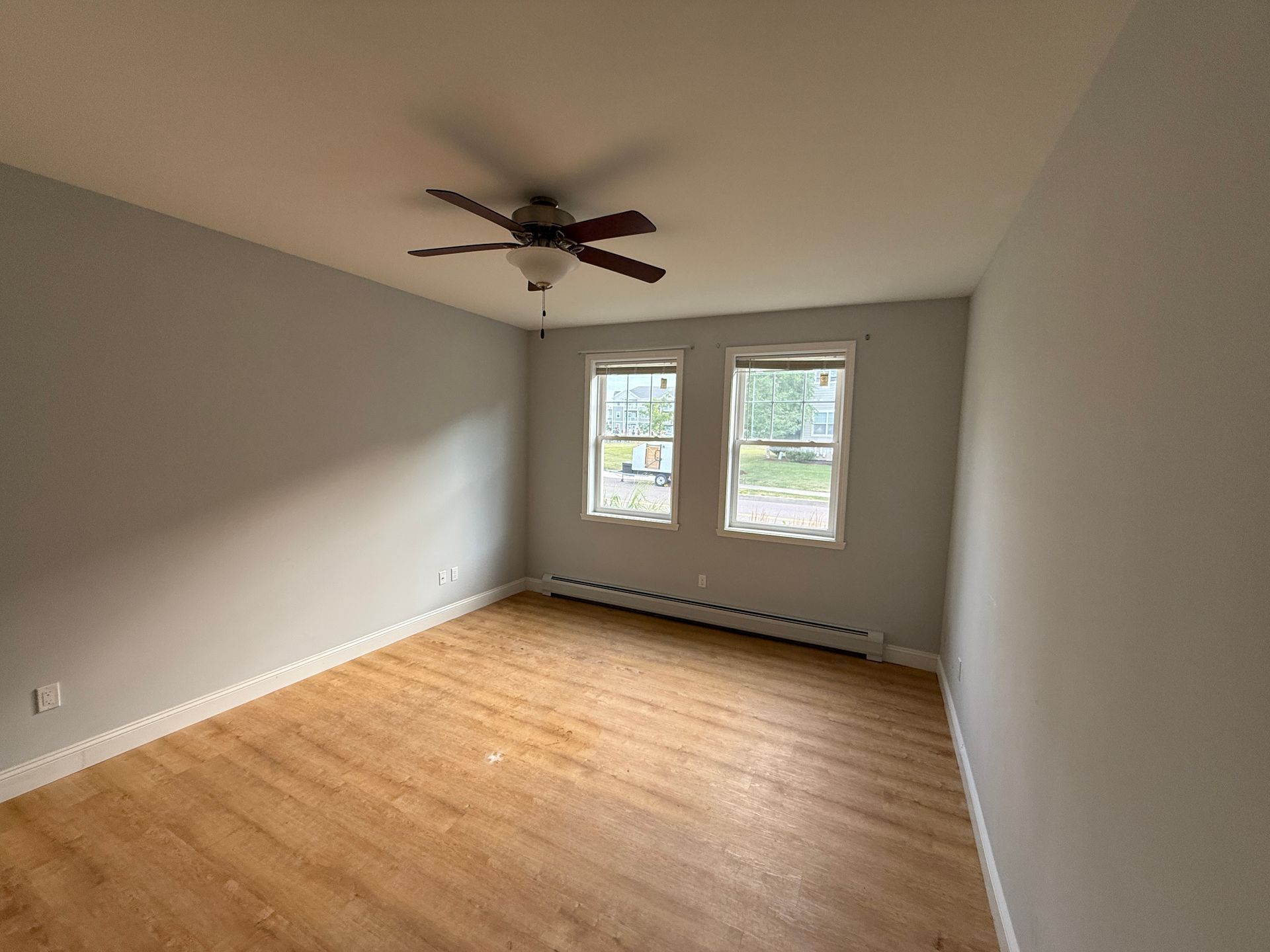 Empty beige bedroom with hardwood floor, ceiling fan, and two windows on the far wall