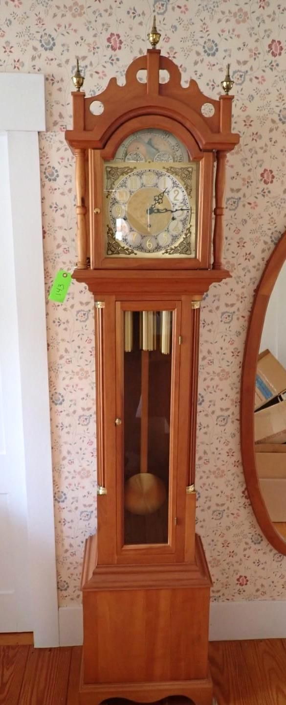 Tall wooden grandfather clock with ornate top and gold clock face against patterned wallpaper.