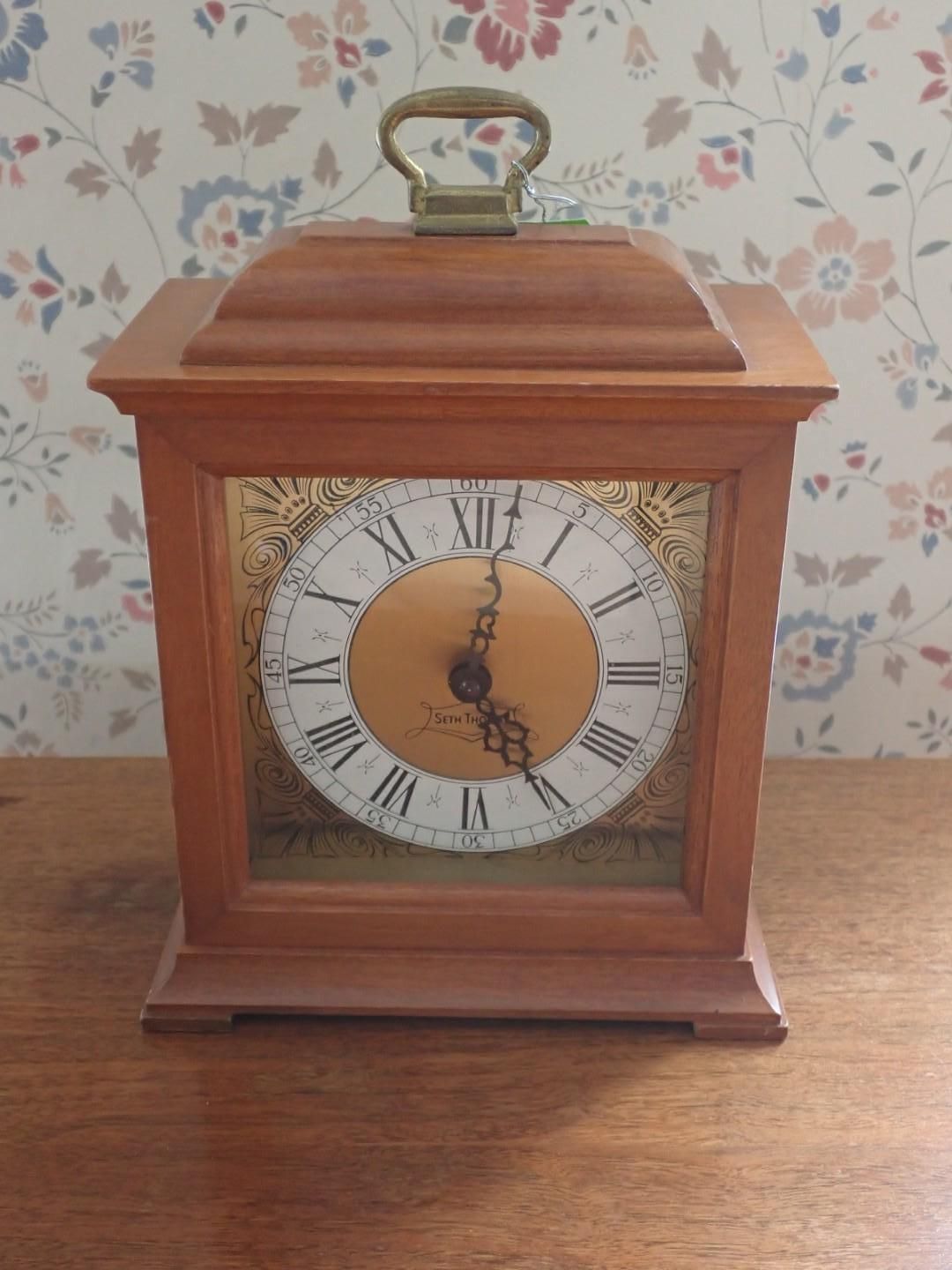 Wooden mantle clock with a white Roman numeral face on a table against floral wallpaper.