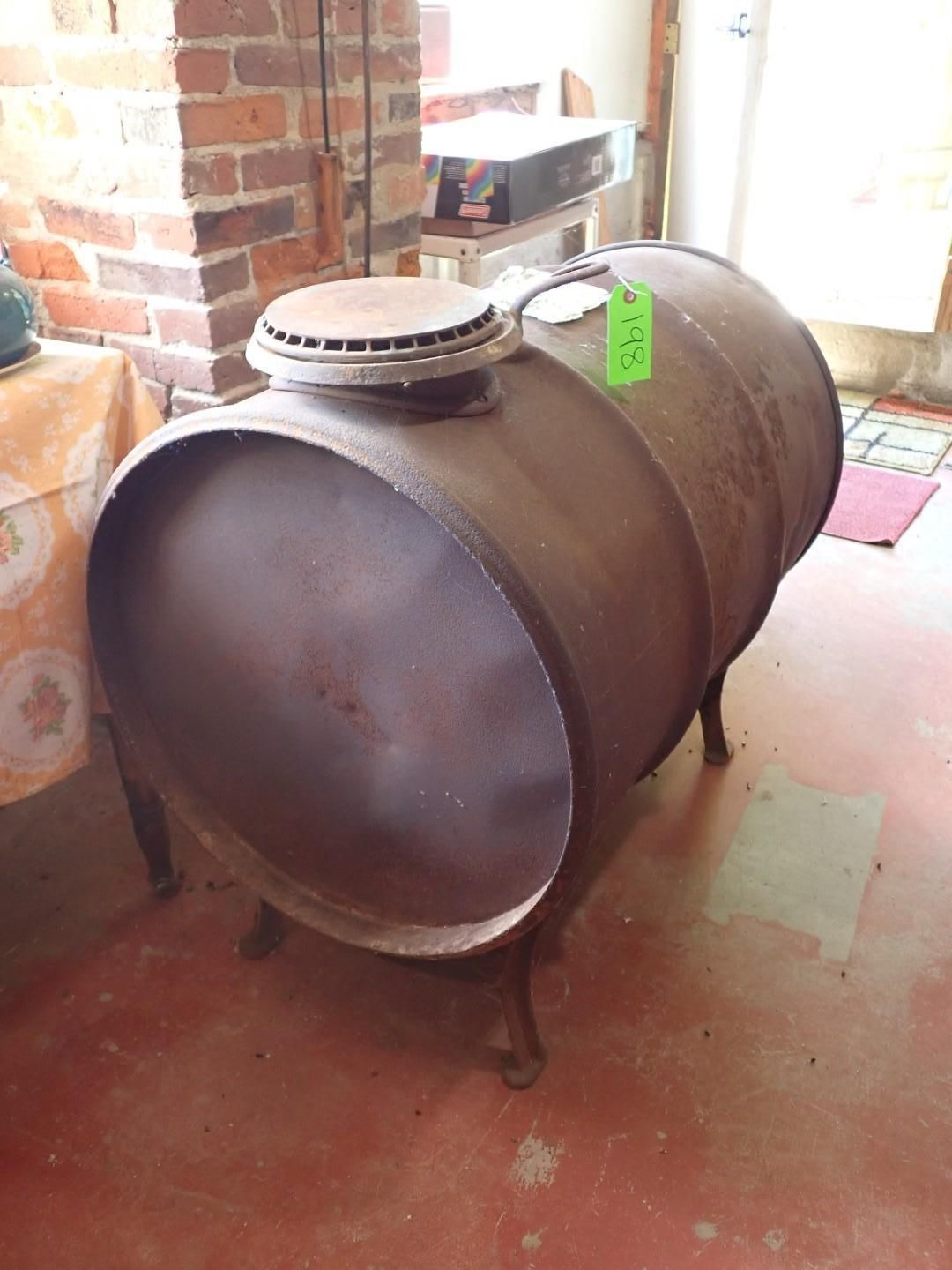 Metal barrel stove with a round lid, sitting indoors beside a brick wall.