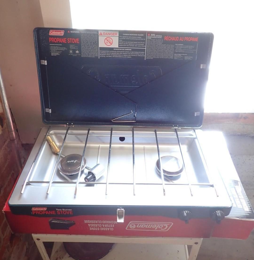 Portable two-burner gas stove with black lid, silver top, and red front panel against a wall