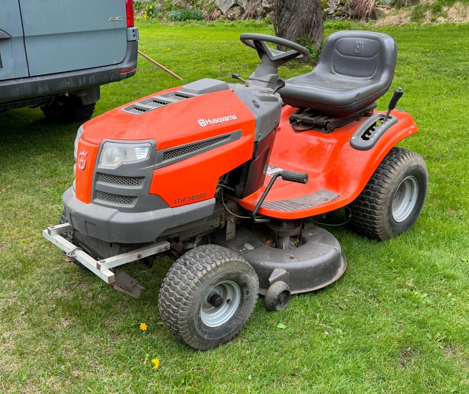 Orange riding lawn mower on grass beside a vehicle, with a black seat and gray cutting deck.