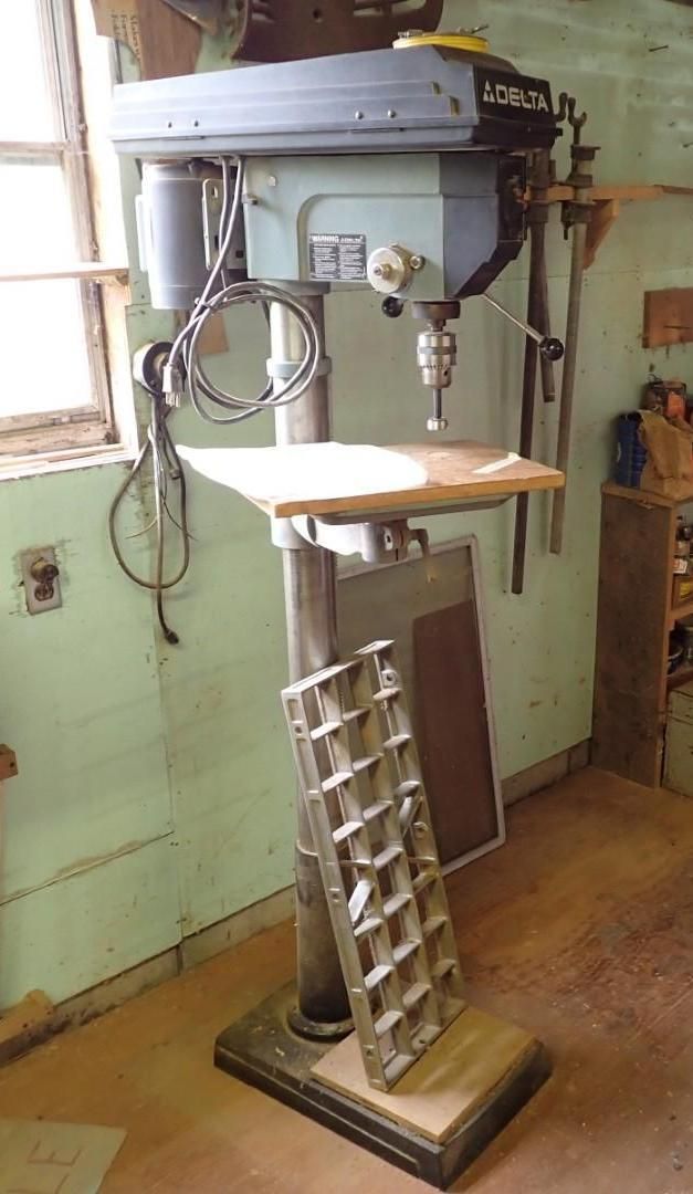 Vintage floor-standing drill press in a workshop with a small wooden worktable and metal base