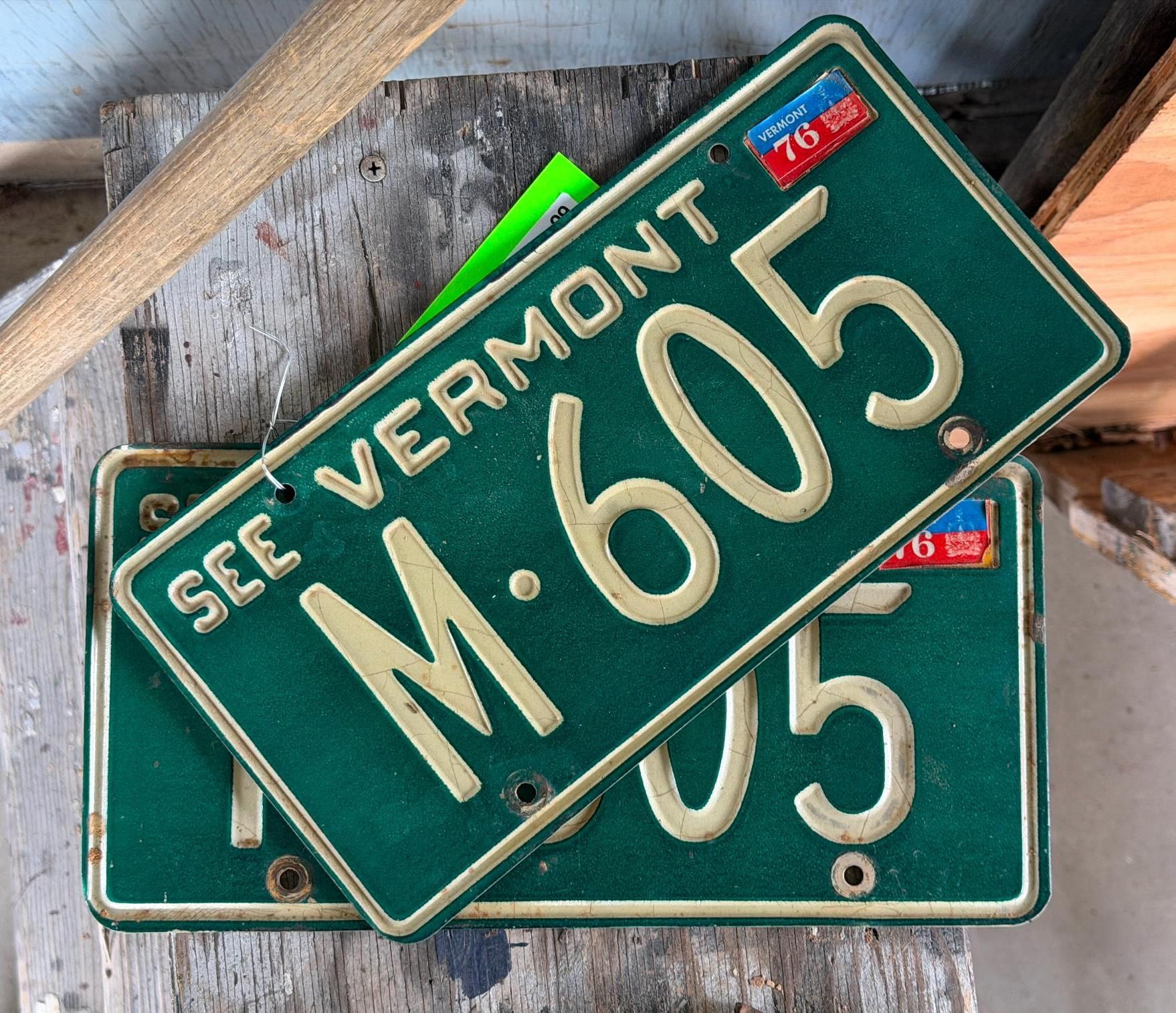 Stacked green Vermont license plates, one reading “SEE VERMONT M-605” on a wooden surface